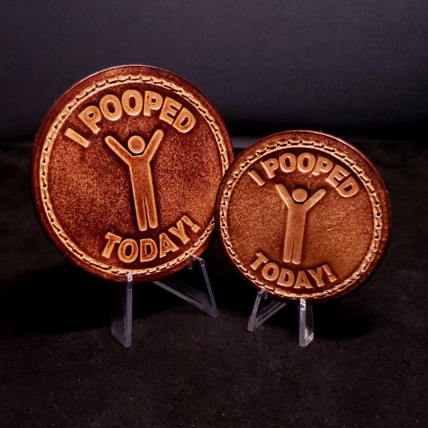 Two brown leather velcro patches embossed with text that says "I pooped today", and a graphic of a cheerful person, posed on a black background.