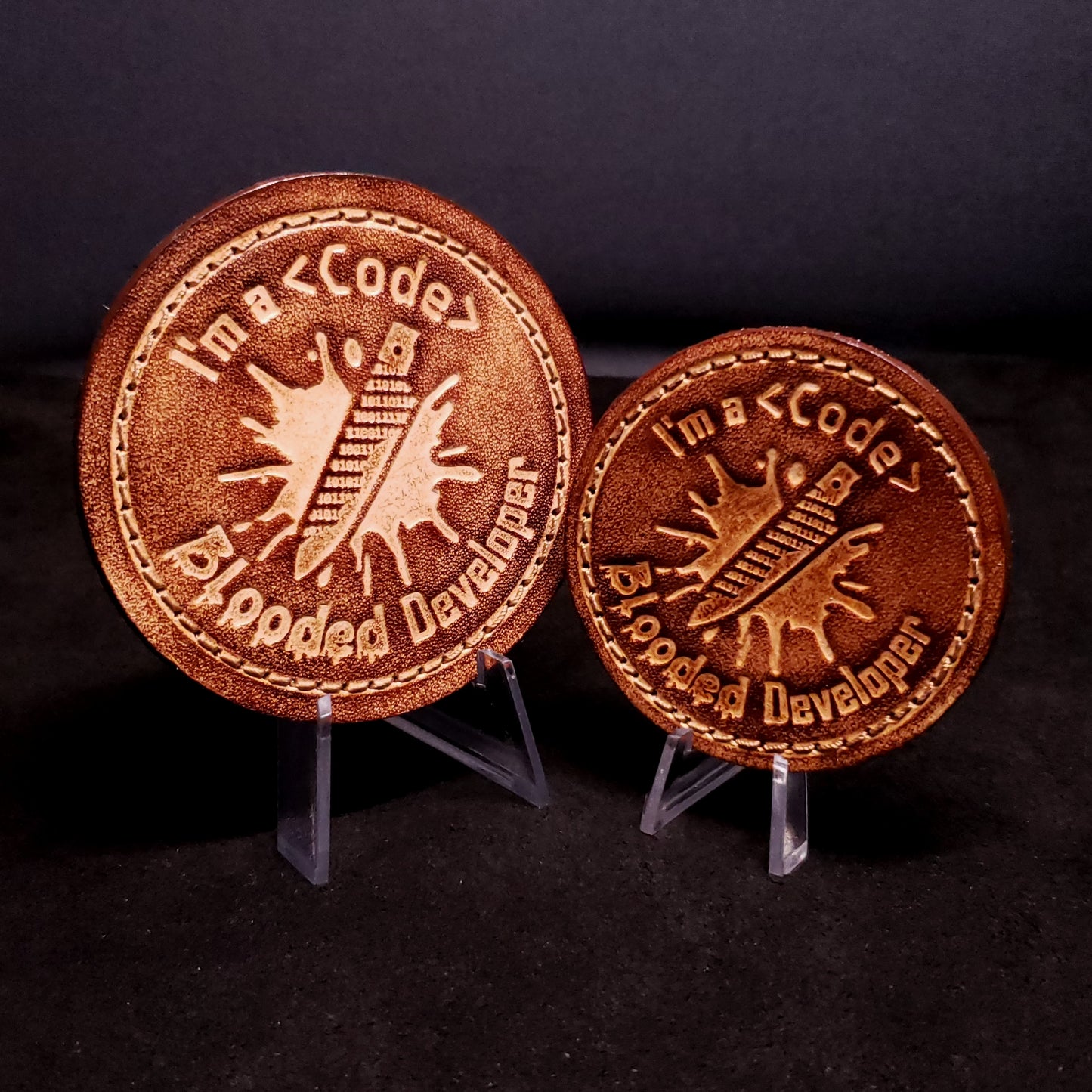 Two brown leatehr velcro patches featuring text and a graphic of a knife made of ones and zeros on a blood splatter, posed on a dark background.