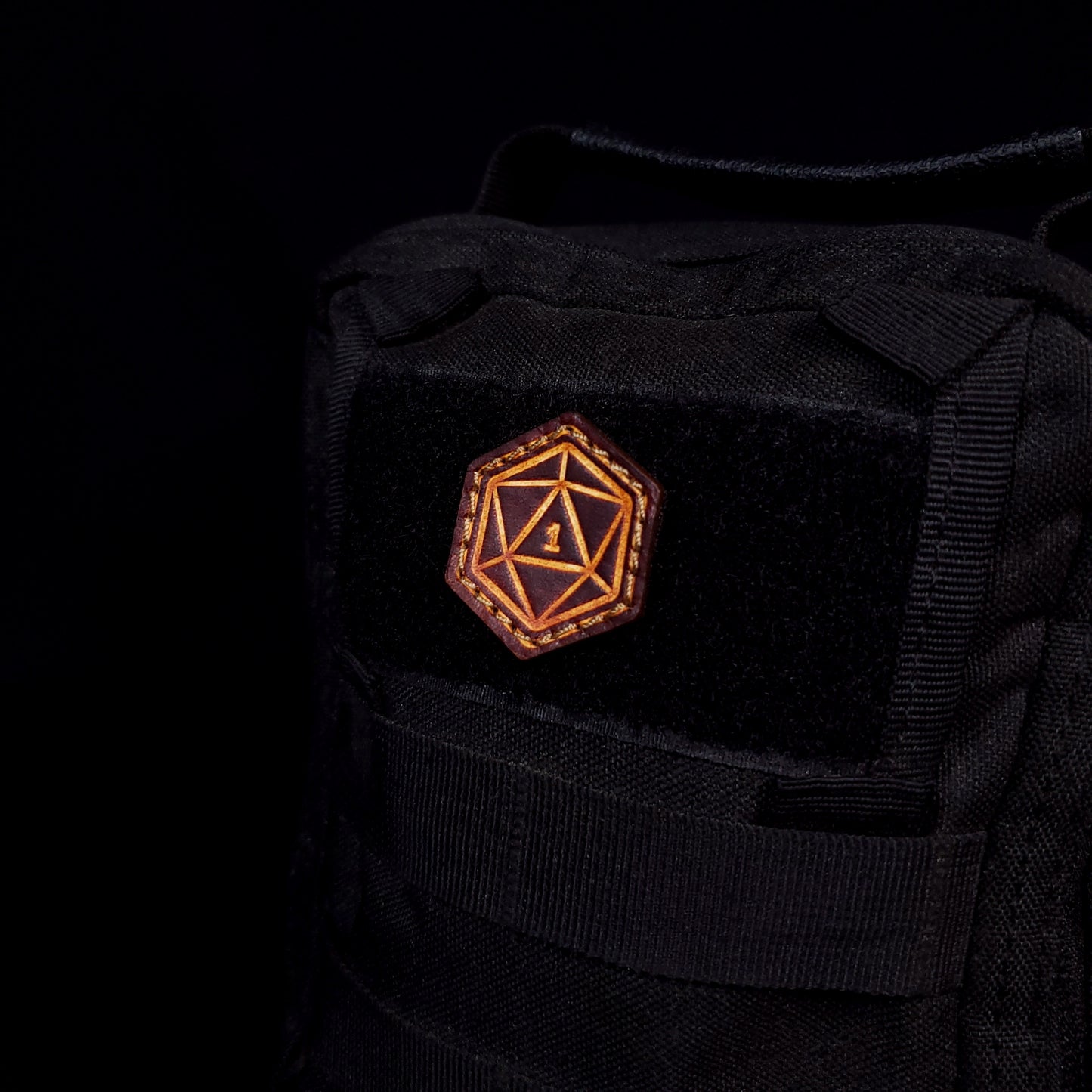 Small leather velcro patch embossed with a twenty-sided die with the number one facing up, affixed to a bag.