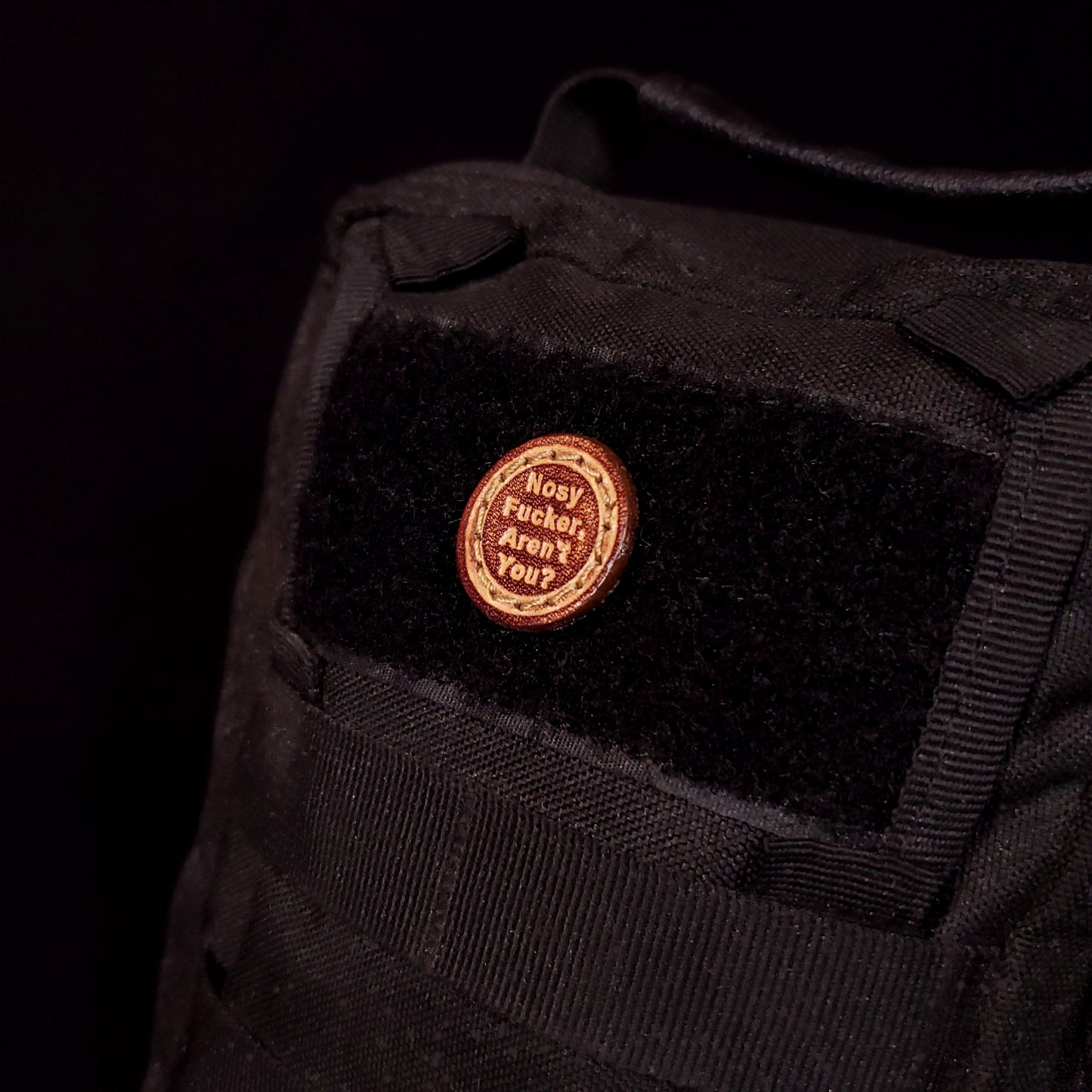 Small leather velcro patch embossed with text that says "Nosy fucker, aren't you?", affixed to a bag.