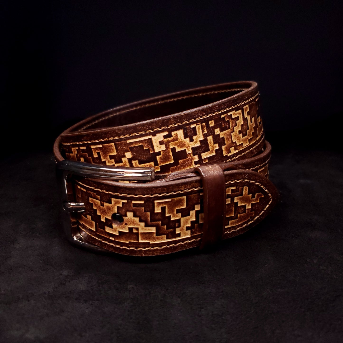 Brown leather belt with a silver buckle, decorated with a digital camouflage pattern and coiled on itself.