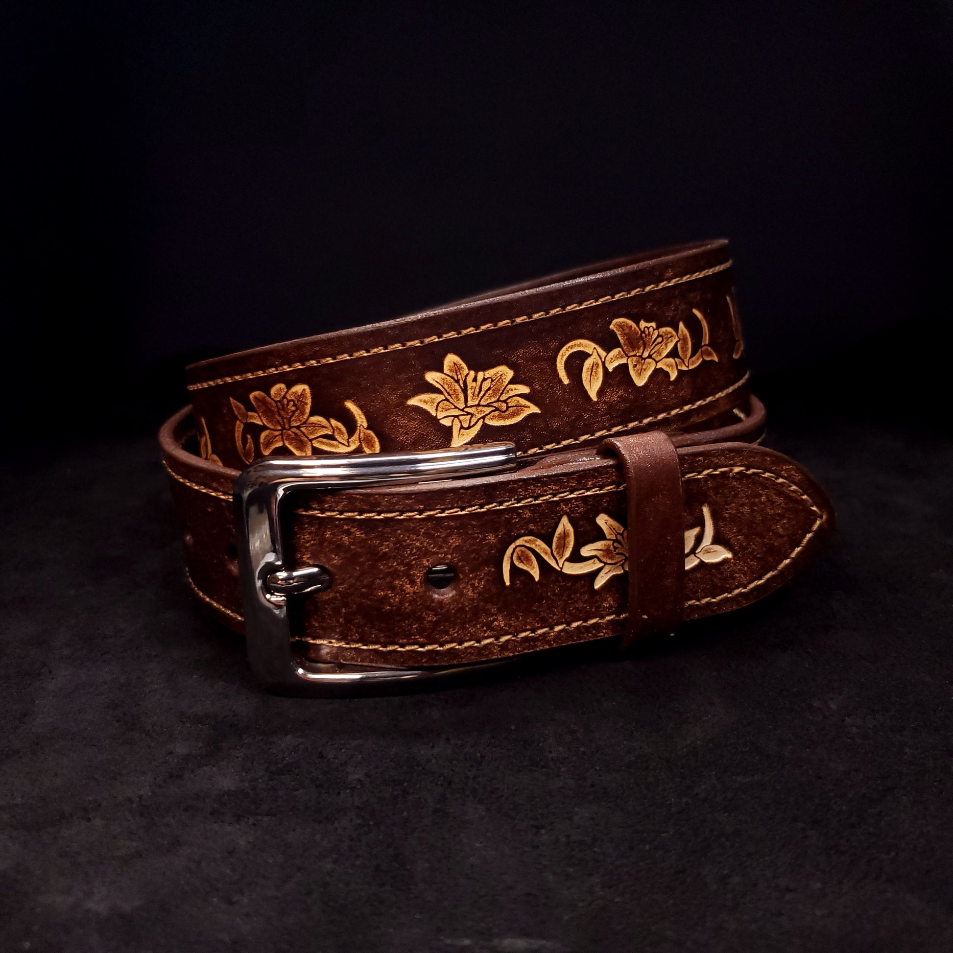 Brown leather belt with a silver buckle, decorated with floral embossing and coiled on itself.