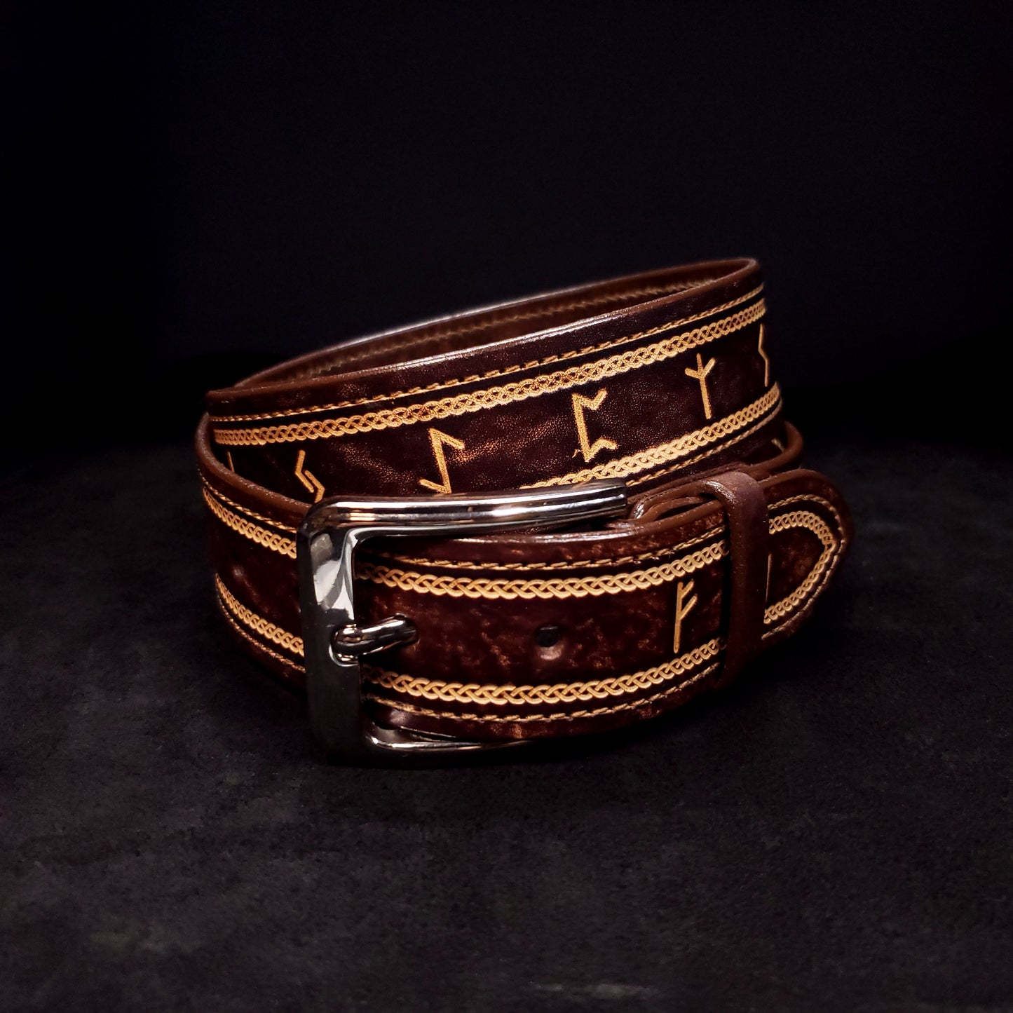 Brown leather belt with a silver buckle, decorated with a Celtic knot pattern and Norse runes, coiled on itself.
