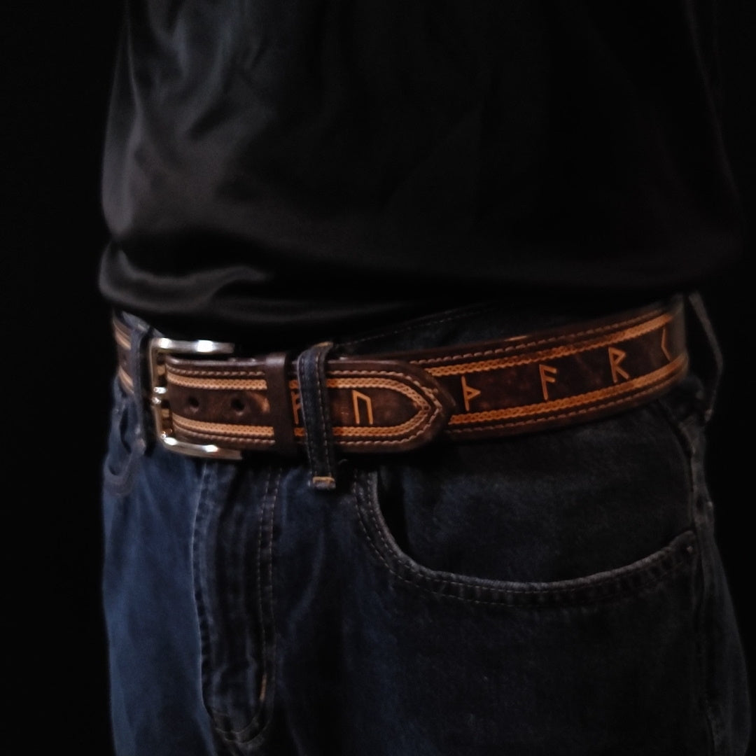 Brown leather belt with a silver buckle and decorated with a Celtic knot pattern and Norse runes, being worn by a man with jeans.