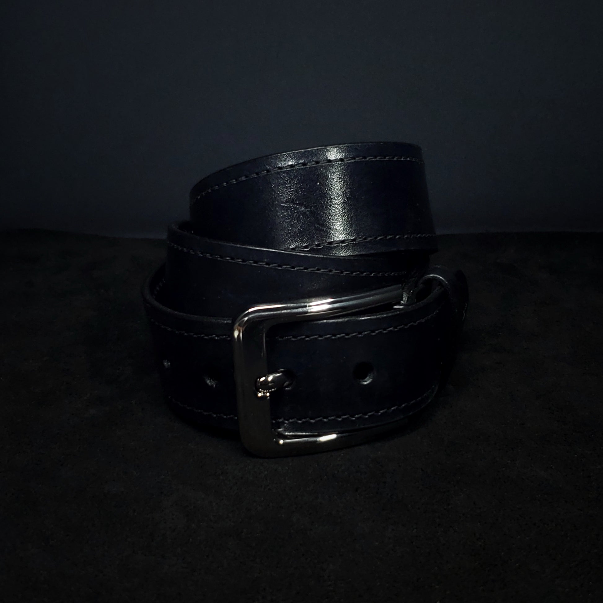 Black leather belt with black stitching and a gun metal buckle, coiled and fastened to its second notch.