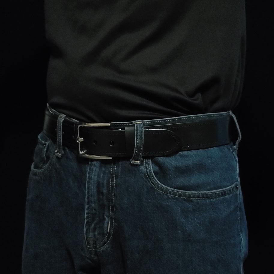 Black leather belt with black stitching and a gun metal buckle, being worn by a man with jeans.