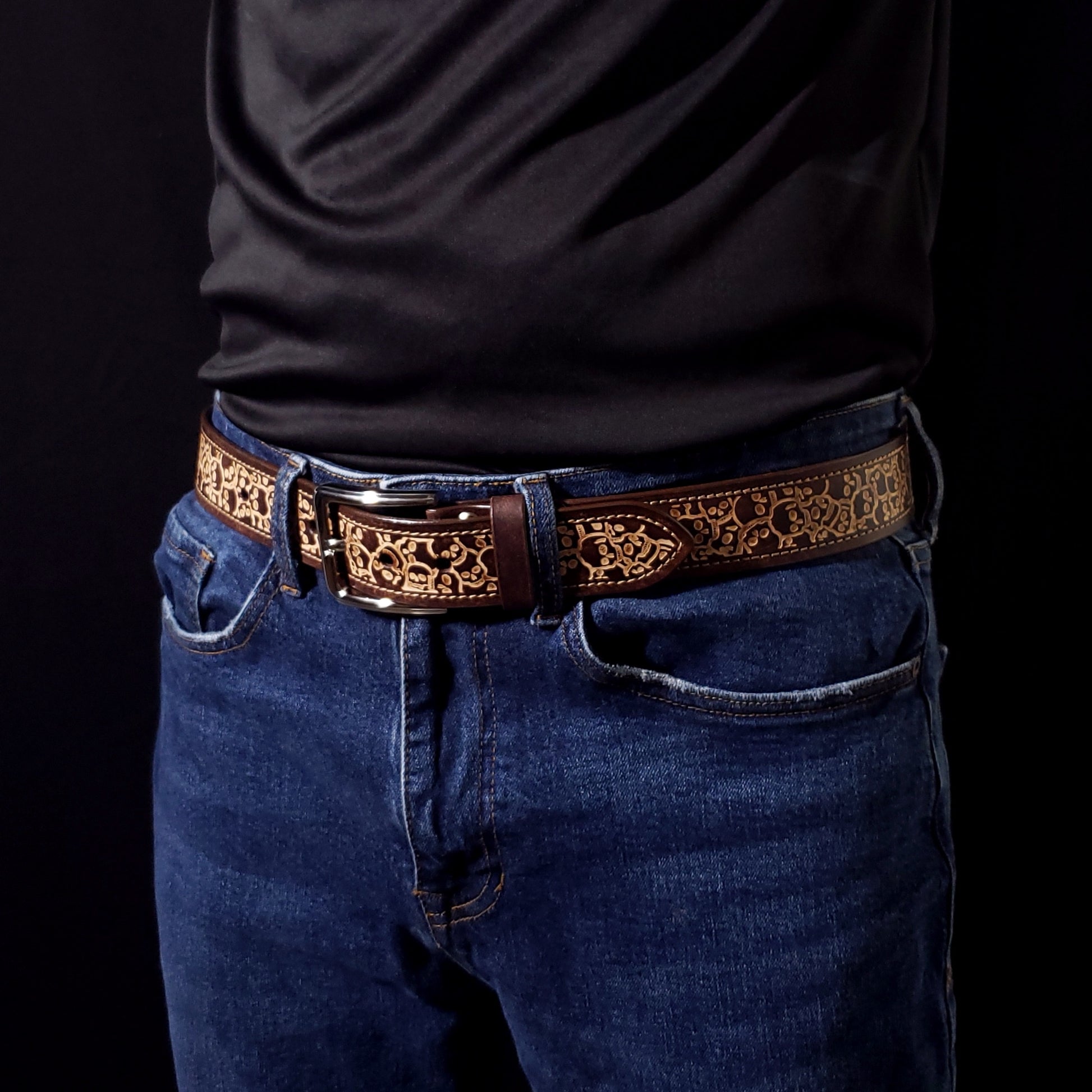 Brown leather belt with a silver buckle and decorated with a skull pattern, being worn by a man with jeans.