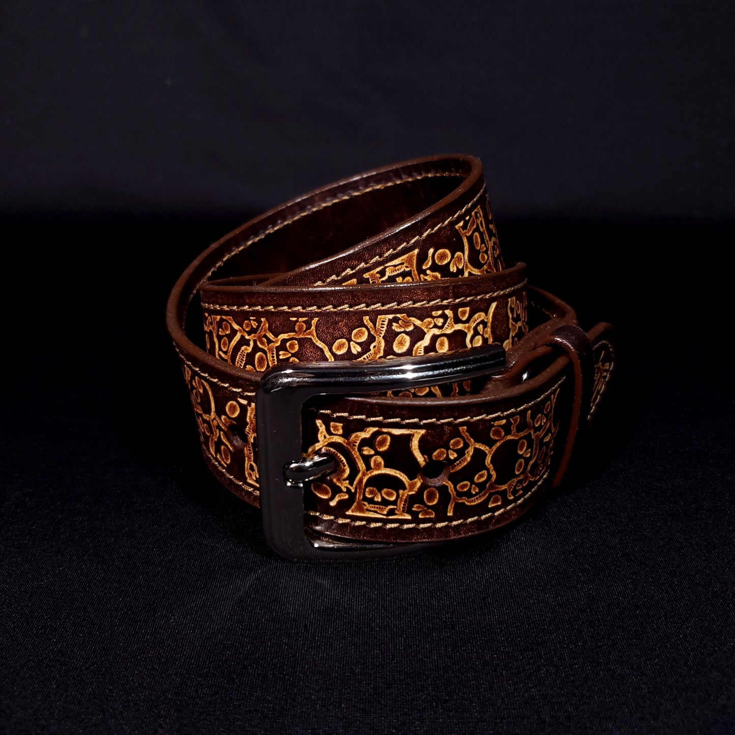 Brown leather belt with a silver buckle, decorated with a skull pattern and coiled on itself.