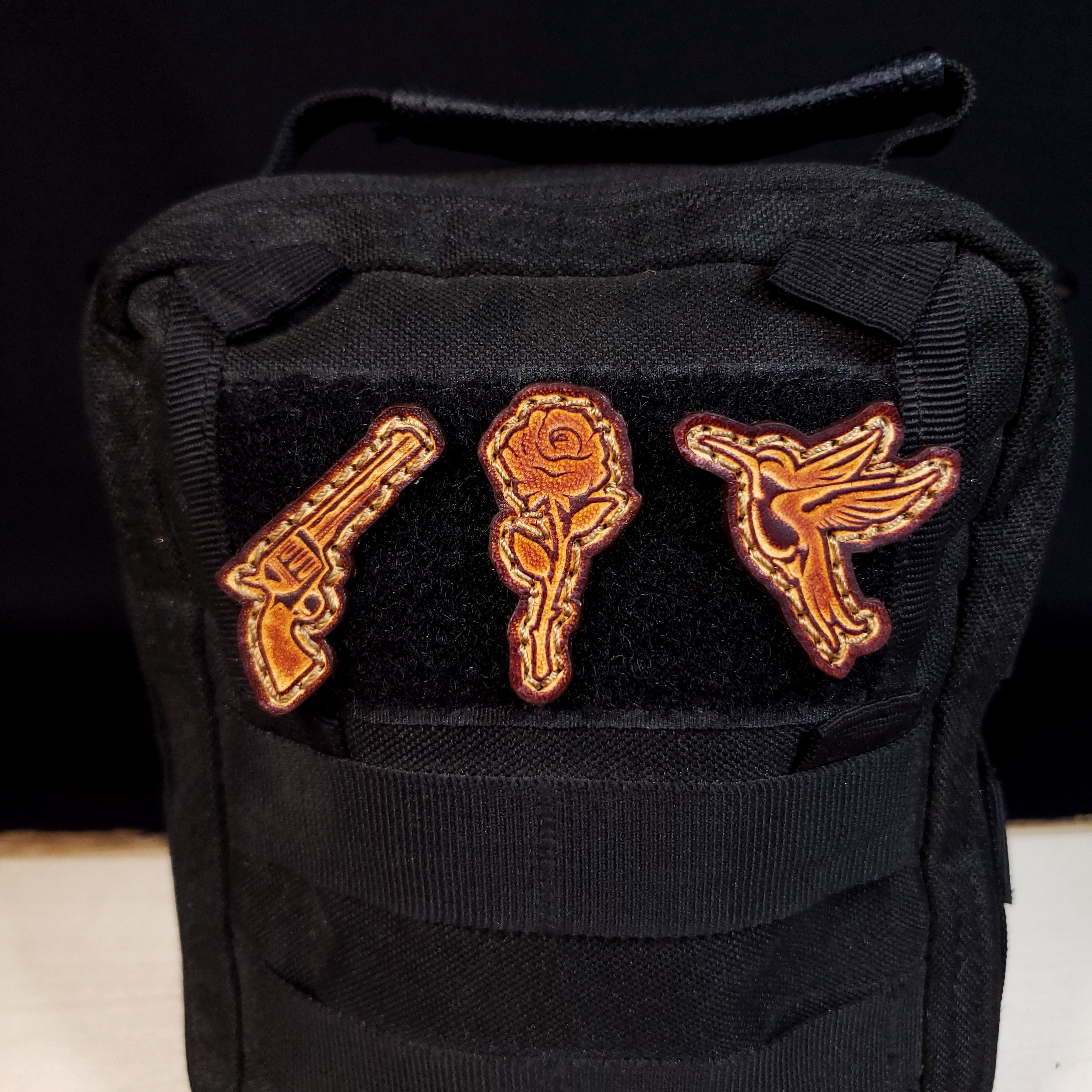 Black pouch with leather patches of a gun, rose, and hummingbird on a dark background