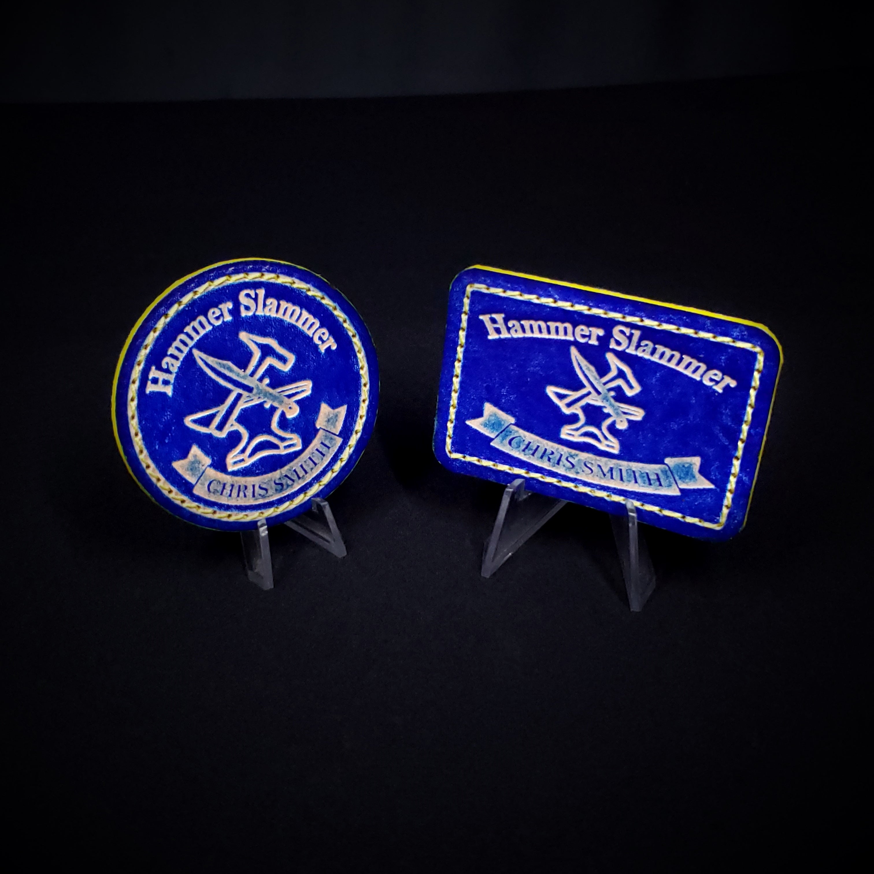Two blue and yellow 'Hammer Slammer' velcro patches on stands against a black background
