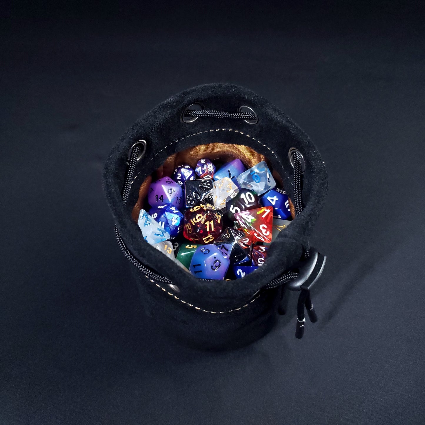 Black suede leather pouch with a brown satin interior and black drawstring, filled with colorful dice.