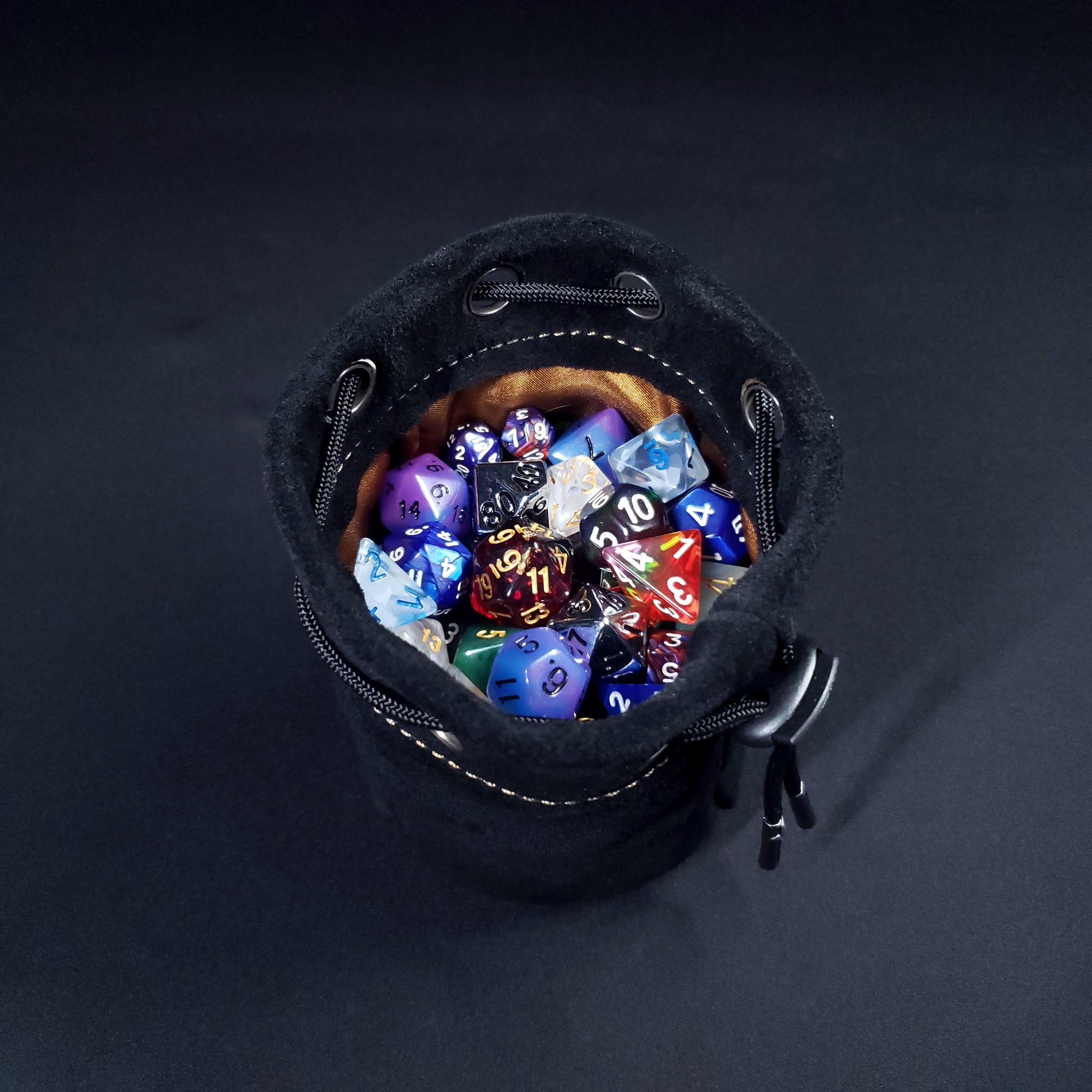 Black suede leather pouch with a brown satin interior and black drawstring, filled with colorful dice.