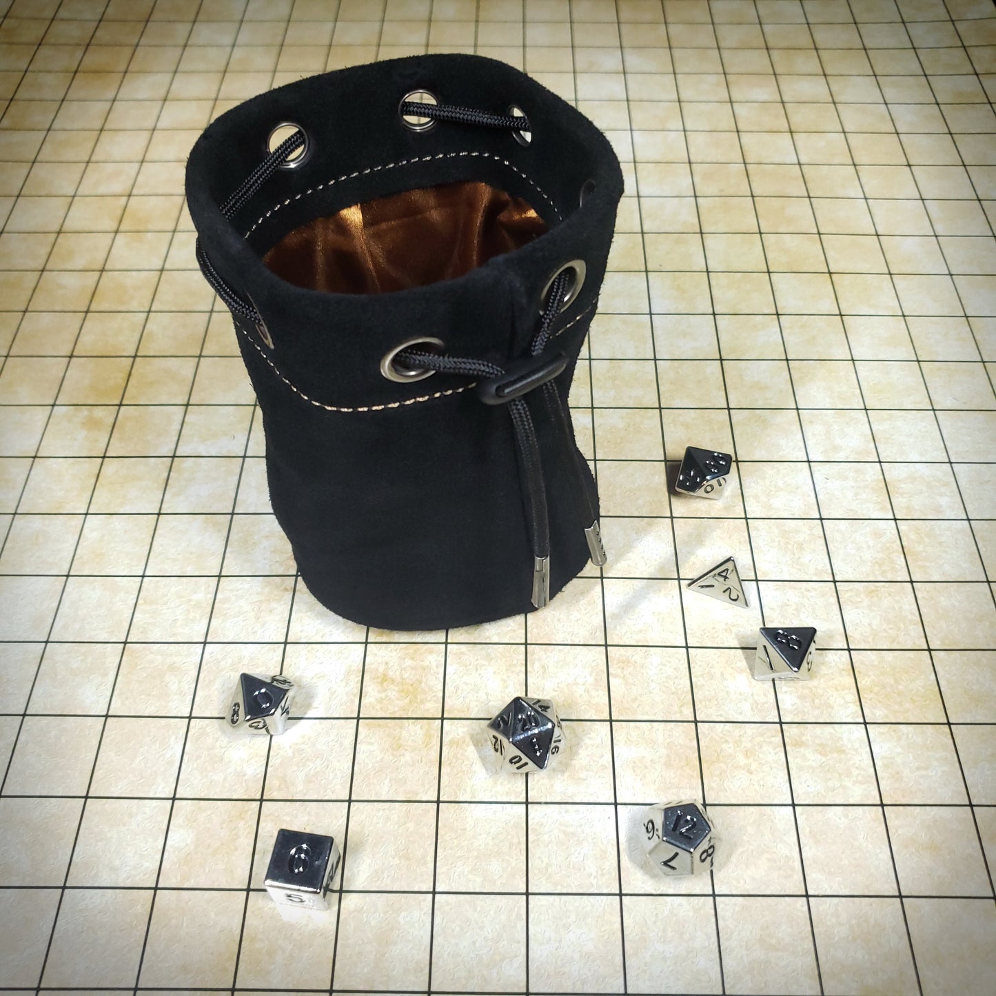 Black suede leather pouch with a brown satin interior and black drawstring, surrounded by seven dice on a battle map grid.