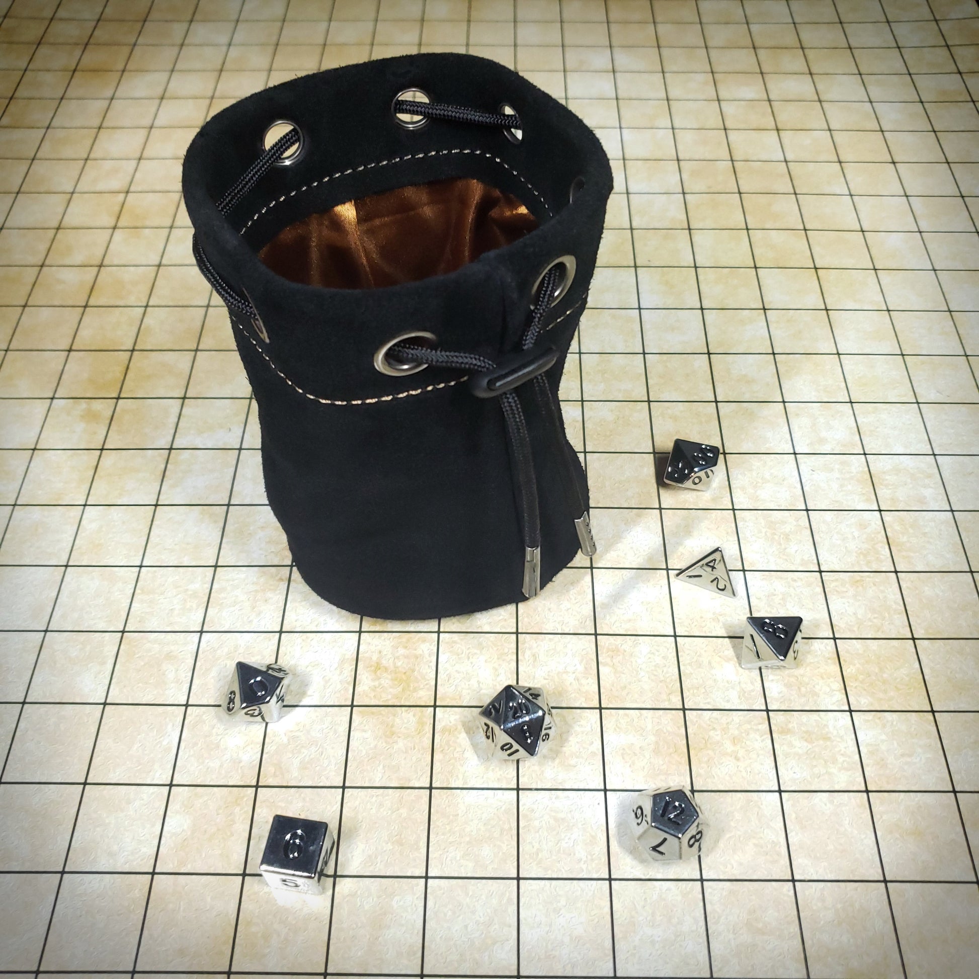 Black suede leather pouch with a brown satin interior and black drawstring, surrounded by seven dice on a battle map grid.