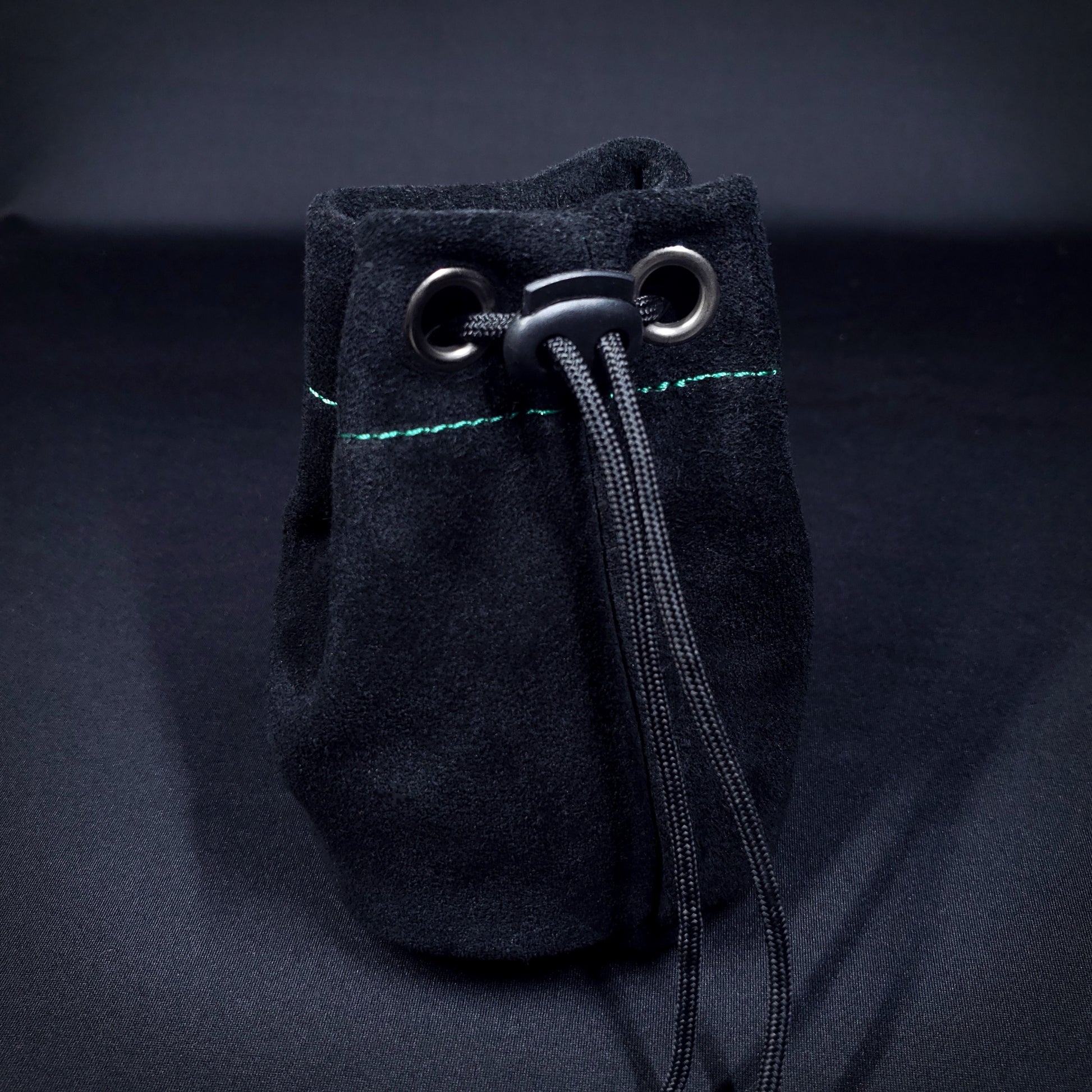 Black suede leather pouch with green stitching and a black drawstring, pulled closed.
