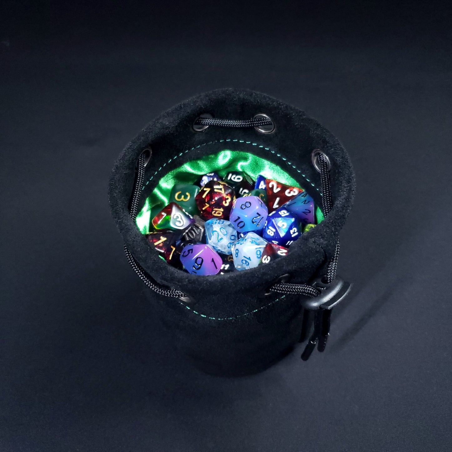 Black suede leather pouch with a green satin interior and black drawstring, filled with colorful dice.