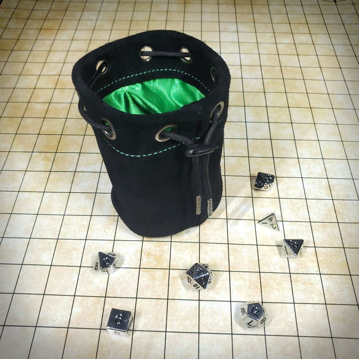 Black suede leather pouch with a green satin interior and black drawstring, surrounded by seven dice on a battle map grid.