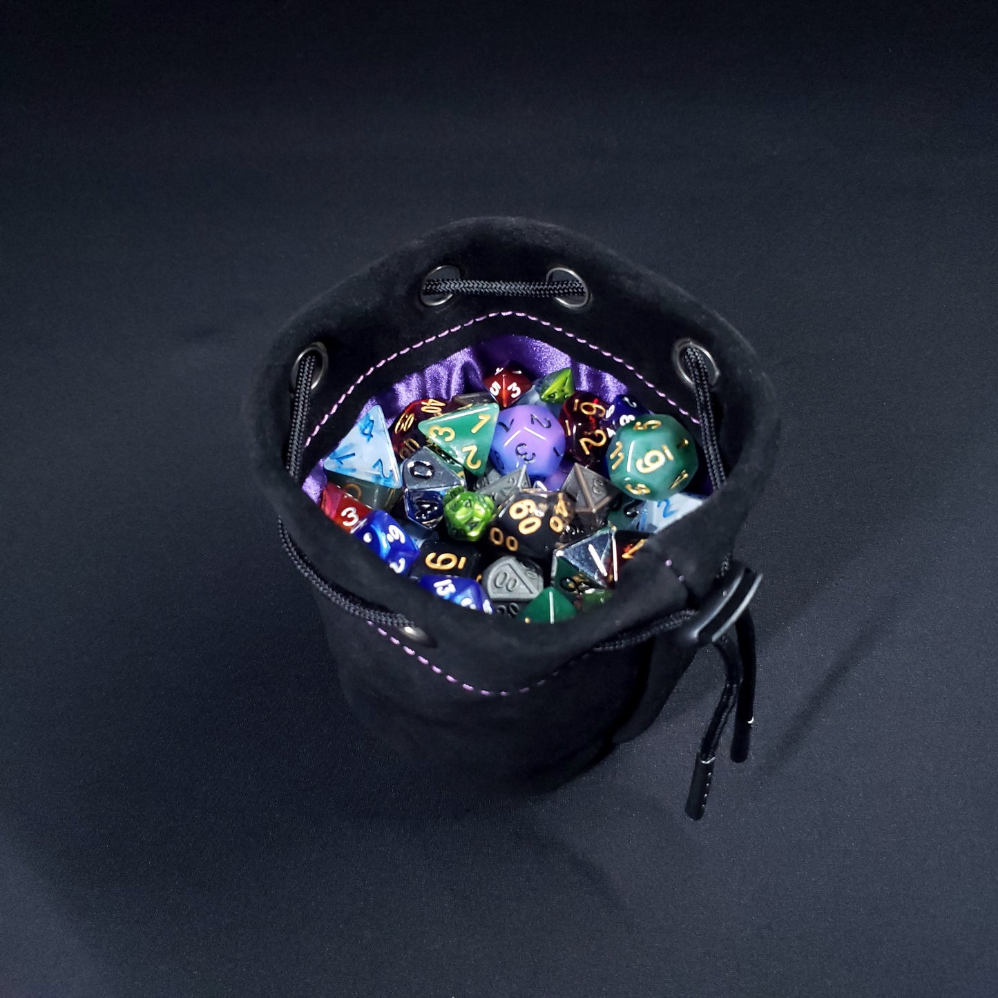 Black suede leather pouch with a purple satin interior and black drawstring, filled with colorful dice.