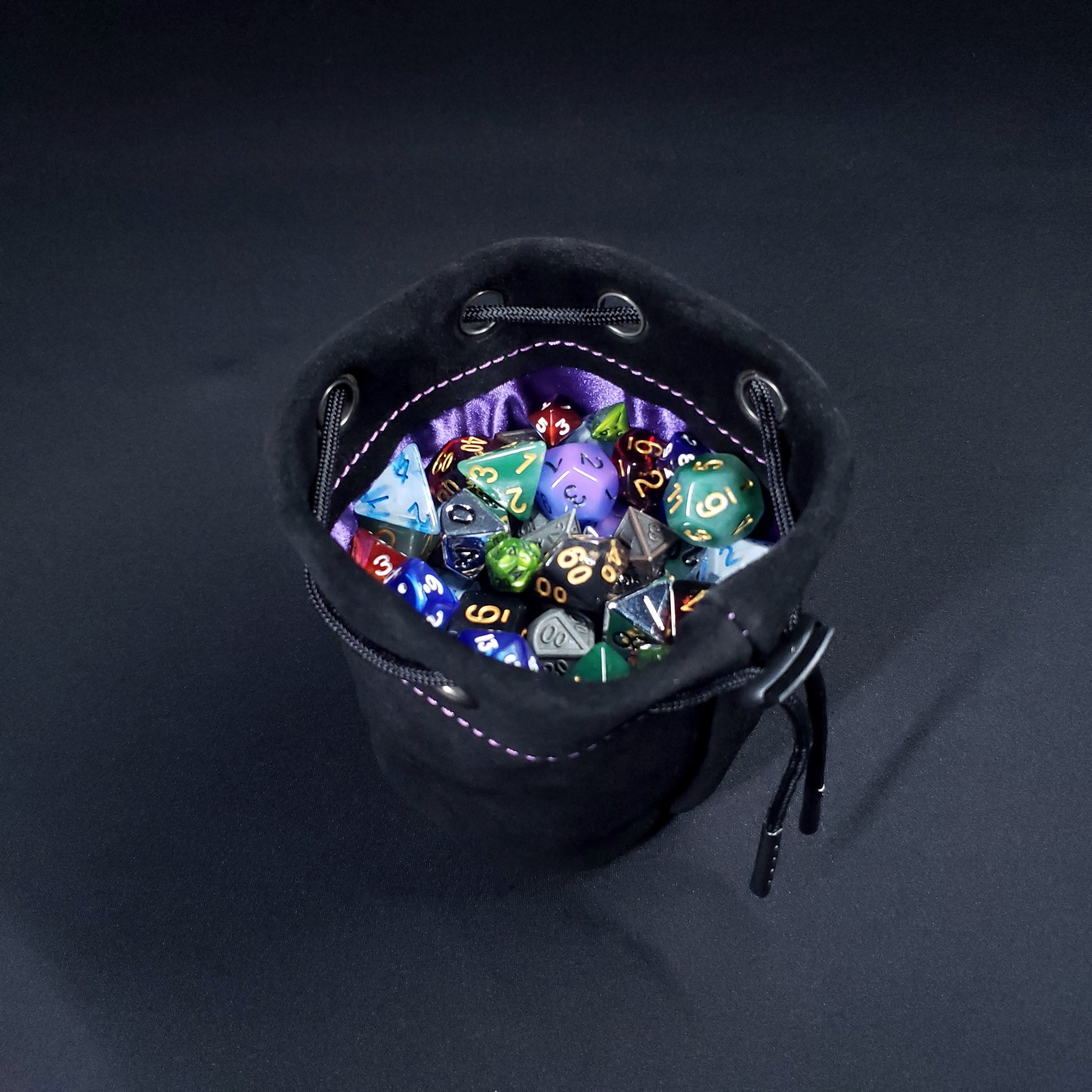 Black suede leather pouch with a purple satin interior and black drawstring, filled with colorful dice.