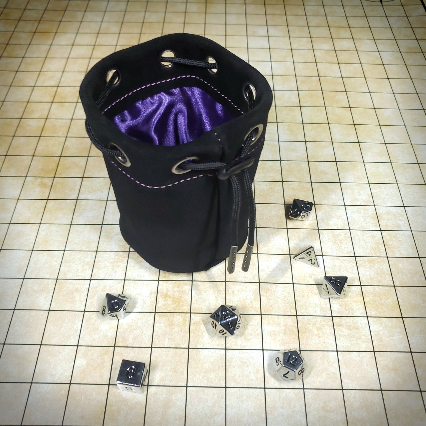 Black suede leather pouch with a purple satin interior and black drawstring, surrounded by seven dice on a battle map grid.