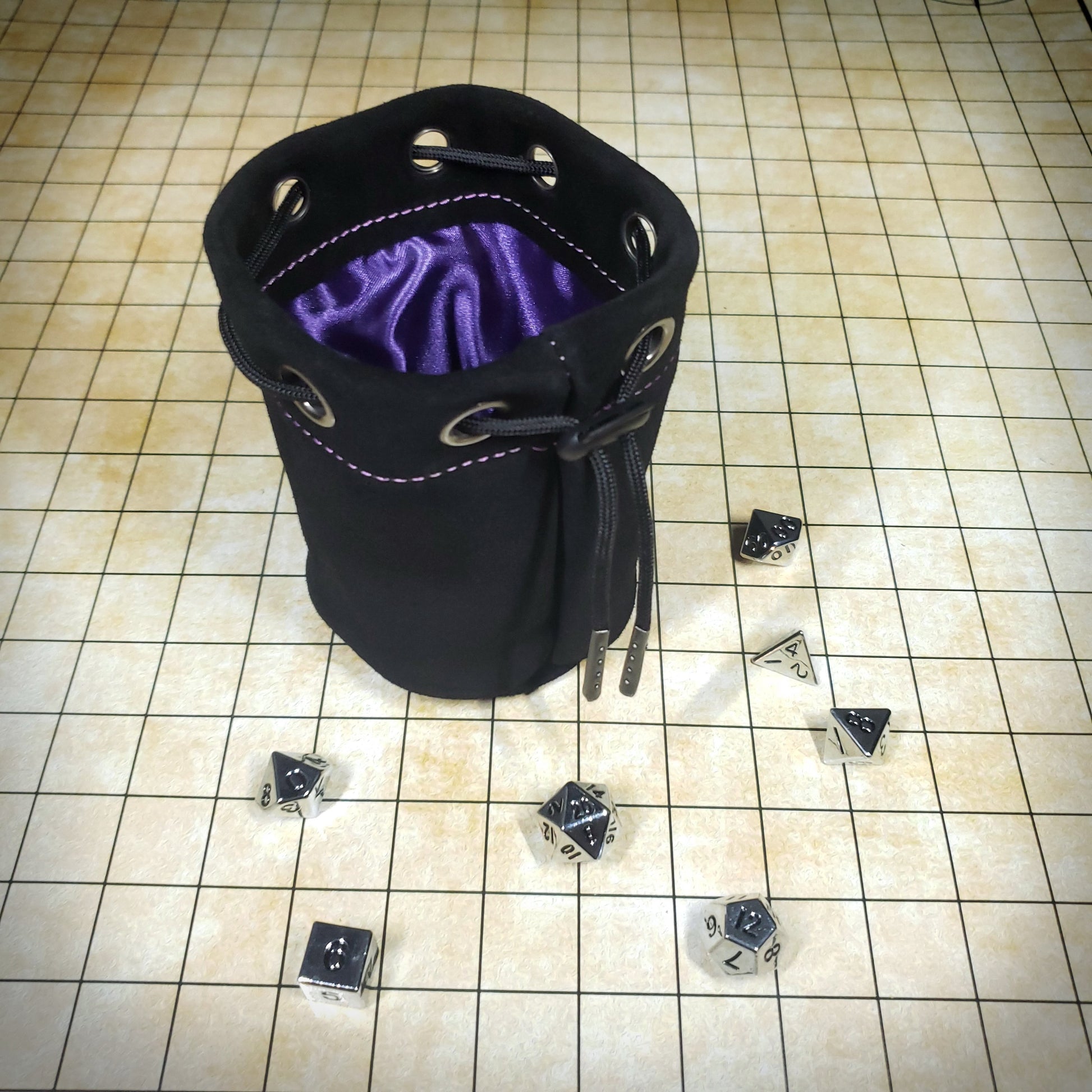 Black suede leather pouch with a purple satin interior and black drawstring, surrounded by seven dice on a battle map grid.