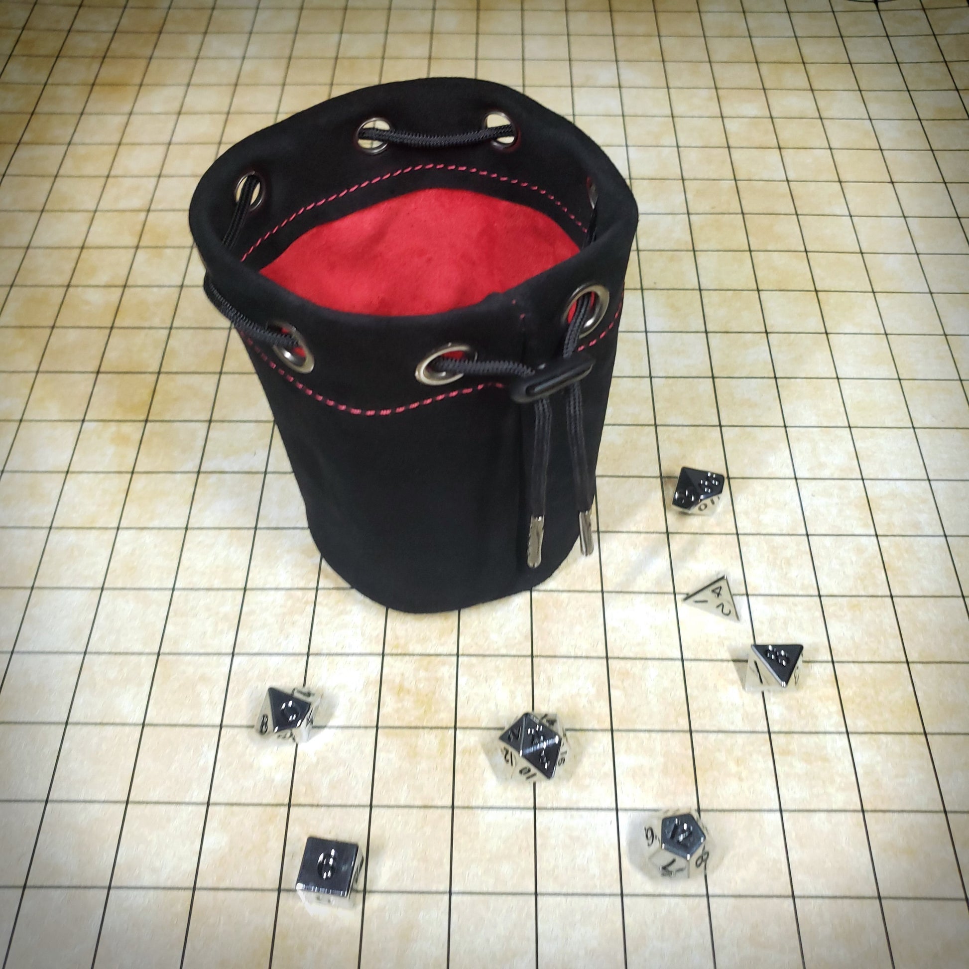 Black suede leather pouch with a red cloth interior and black drawstring, surrounded by seven dice on a battle map grid.