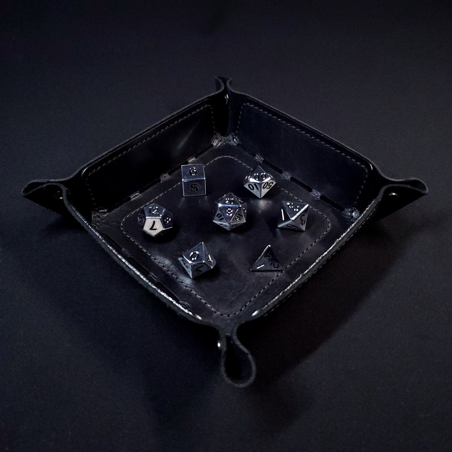 Black leather dice tray with black stitching and gun metal buttons with seven dice inside it.