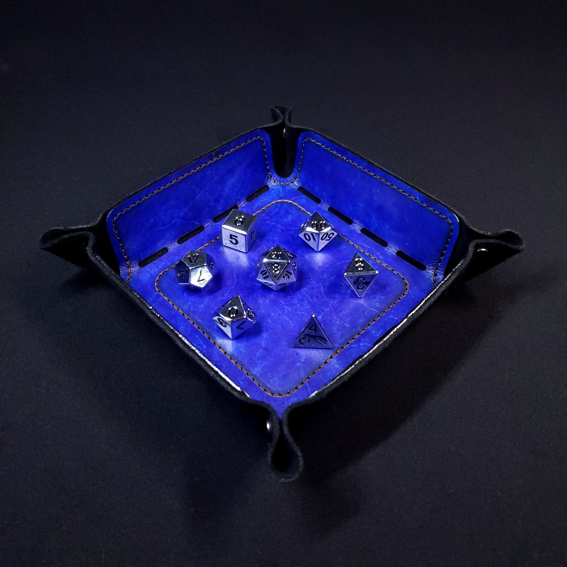 Blue leather dice tray with black stitching and gun-metal buttons with seven dice inside it.