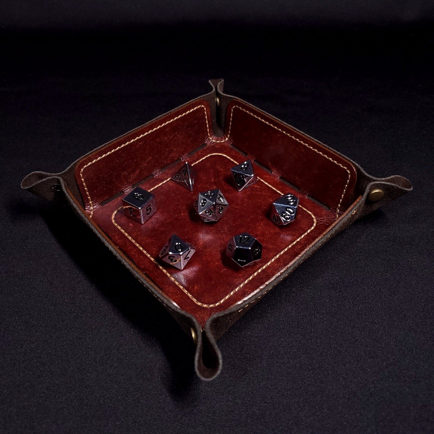Brown leather dice tray with light brown stitching and bronze buttons with seven dice inside it.