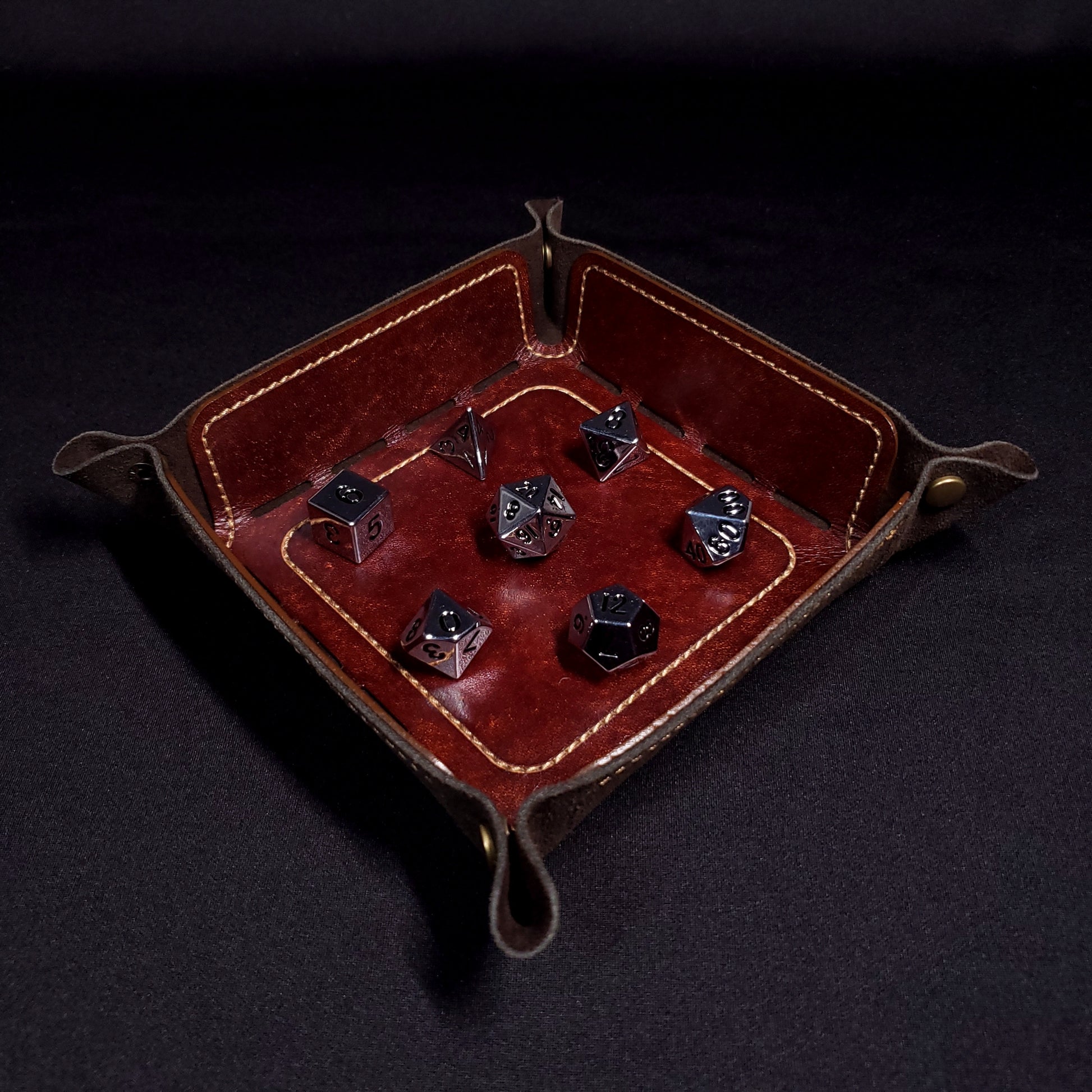 Brown leather dice tray with light brown stitching and bronze buttons with seven dice inside it.