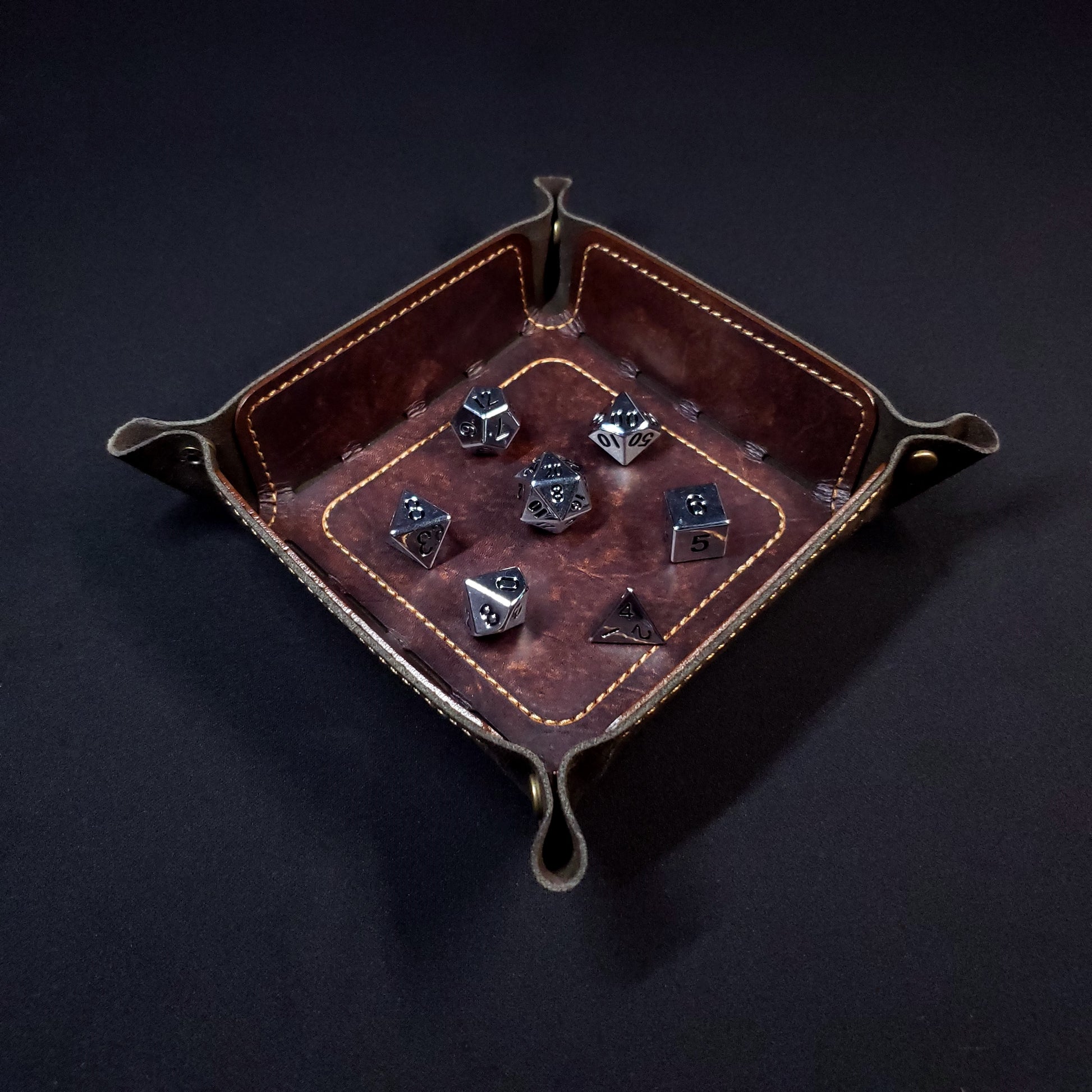 Brown leather dice tray with light brown stitching and bronze buttons with seven dice inside it.