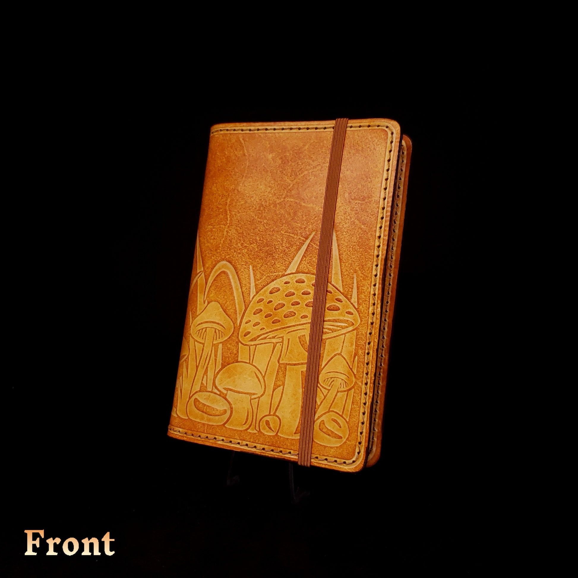 Brown leather notebook cover that's embossed with a mushroom design and closed with an elastic strap.