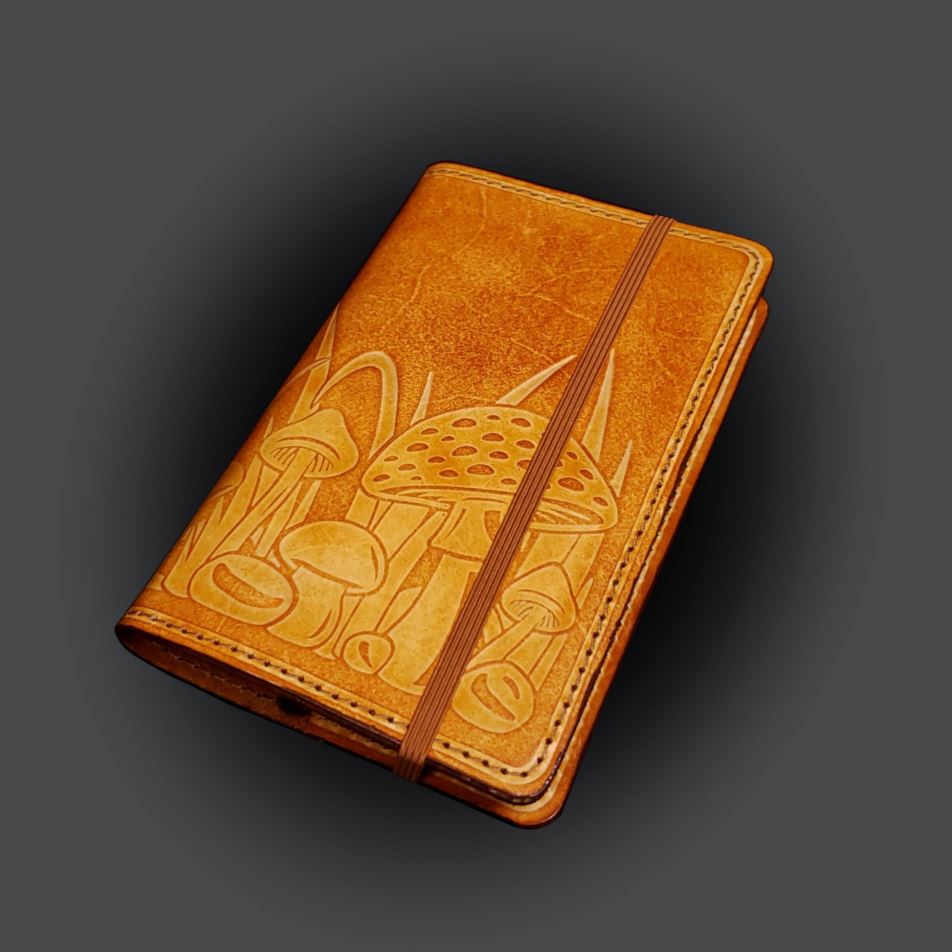 Brown leather notebook cover that's embossed with a mushroom design and closed with an elastic strap.