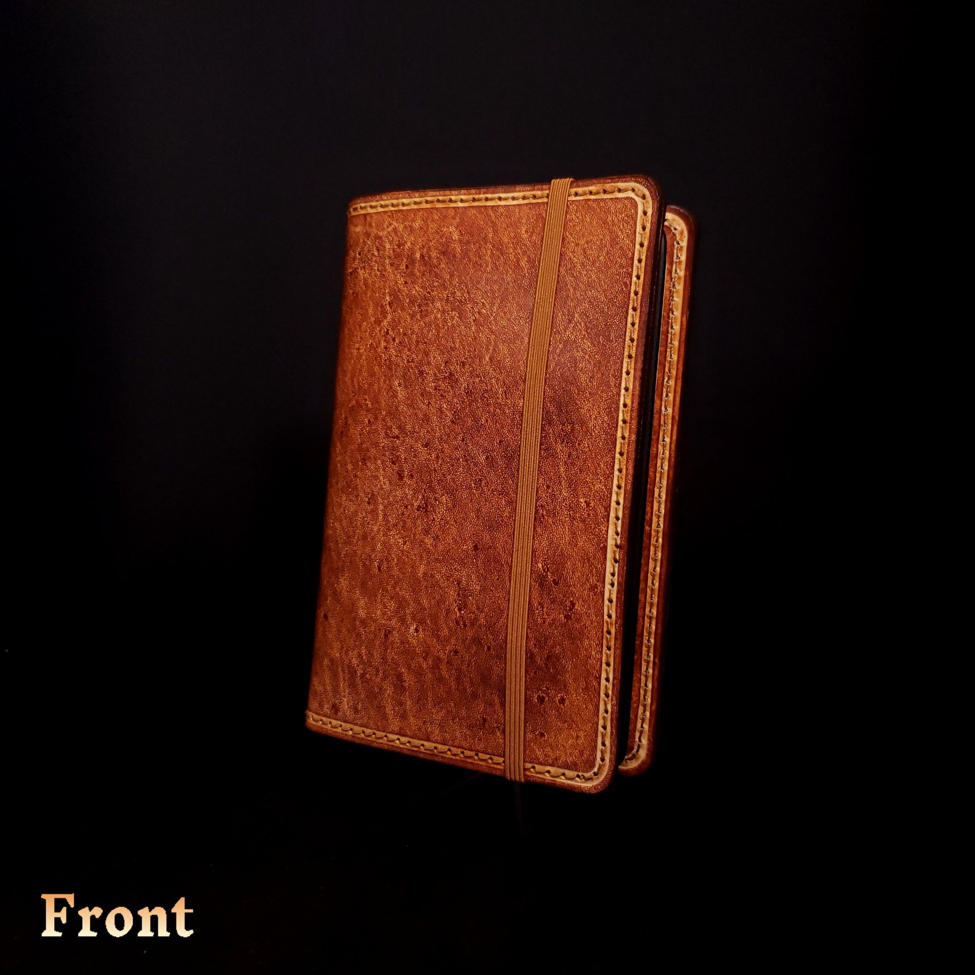 Brown leather notebook cover, closed with an elastic strap, viewed from the front.