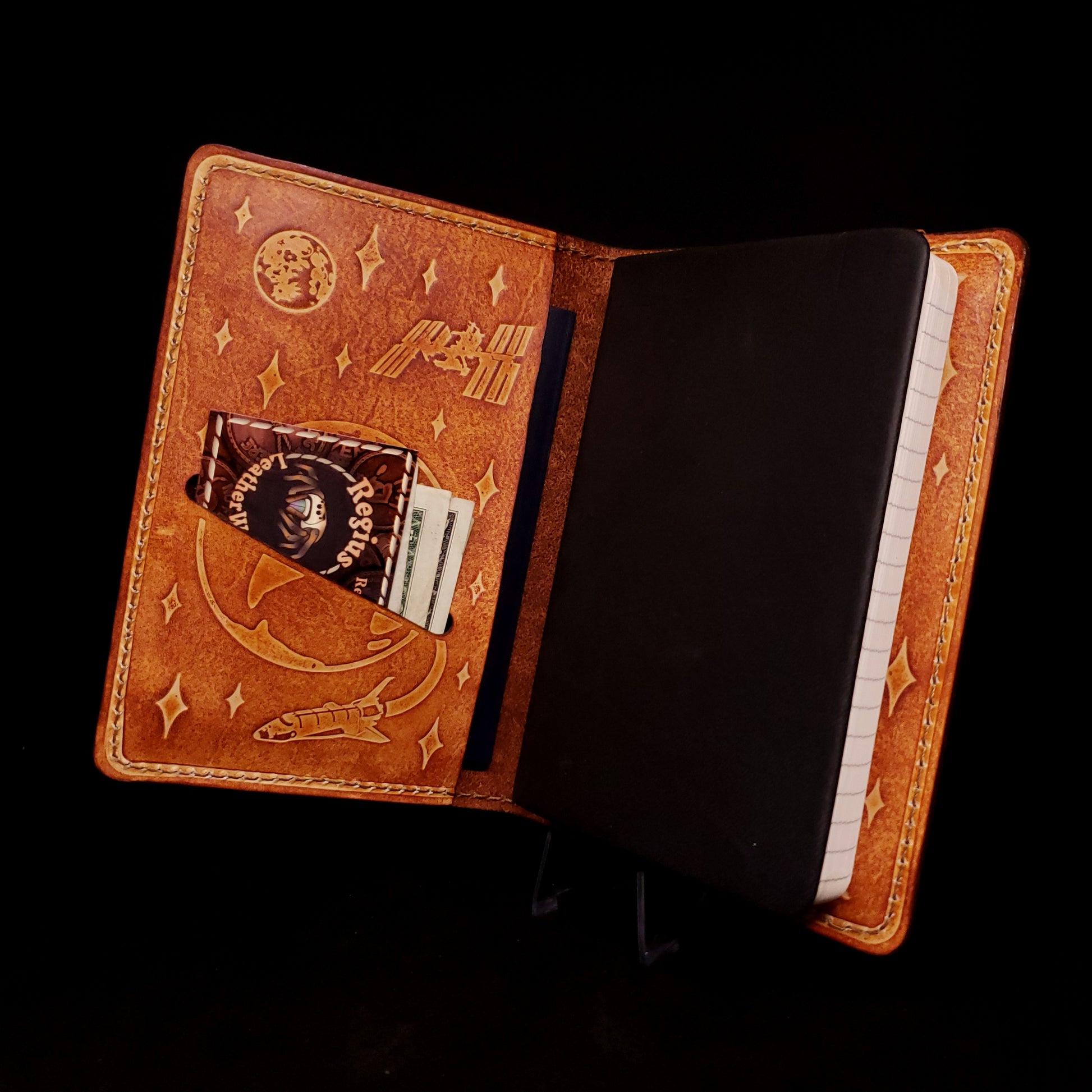 Brown leather notebook cover holding a pocket journal, embossed with a satellite and space shuttle flying around earth and the moon.