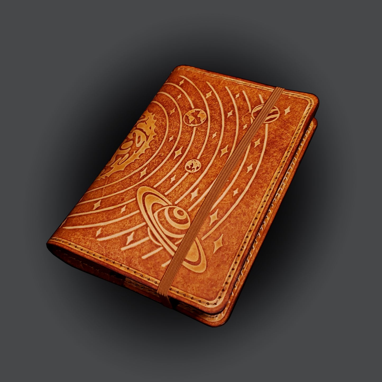 Brown leather notebook cover that's embossed with the solar system, closed with an elastic strap.