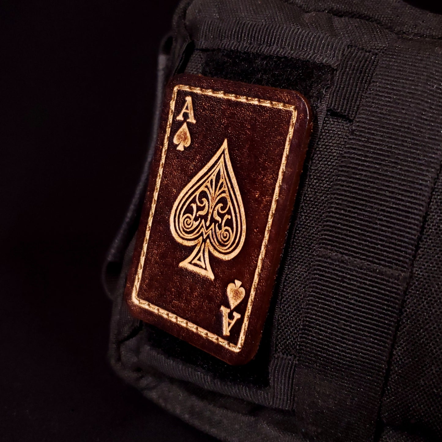 Brown rectangular velcro patch embossed to resemble an ace of spades playing card, affixed to a bag.