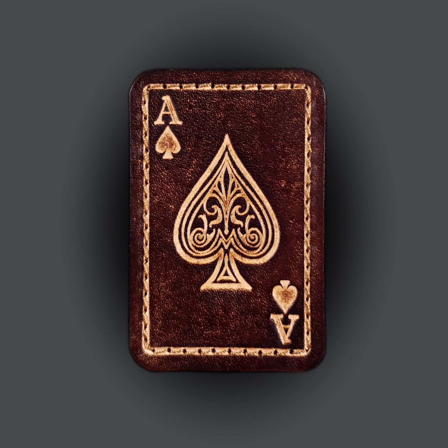 Brown rectangular velcro patch embossed with graphics and letters to resemble an ace of spades playing card.