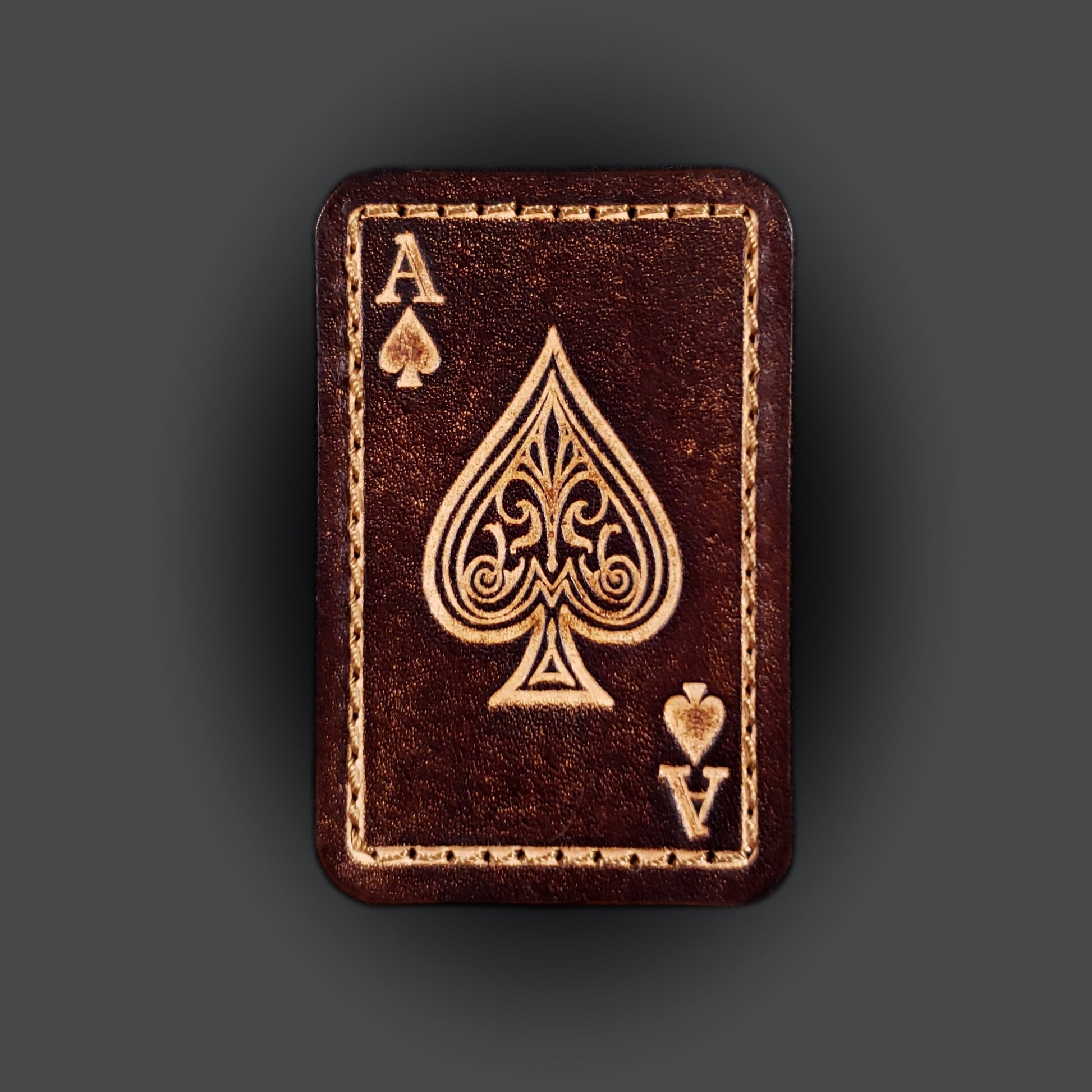 Brown rectangular velcro patch embossed with graphics and letters to resemble an ace of spades playing card.