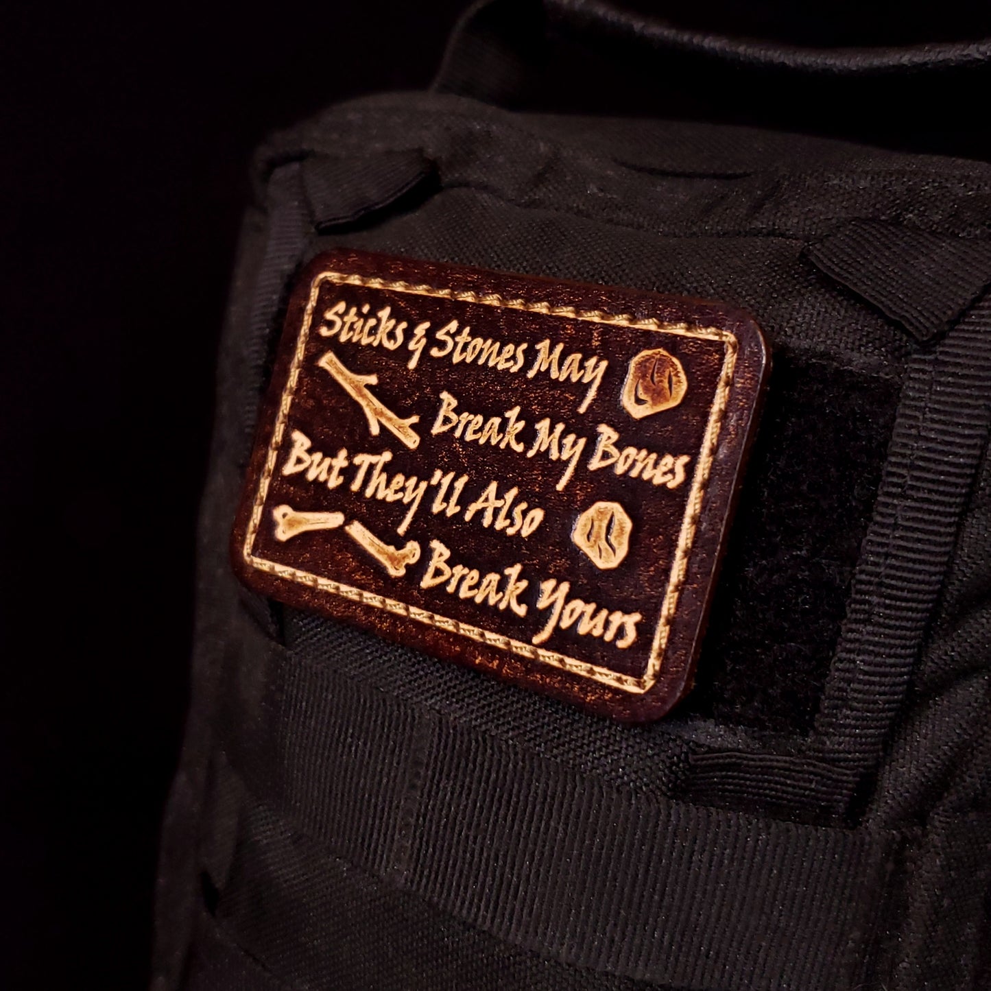 Brown rectangular velcro patch embossed with text and graphics of sticks, rocks, and a broken bone, affixed to a bag.