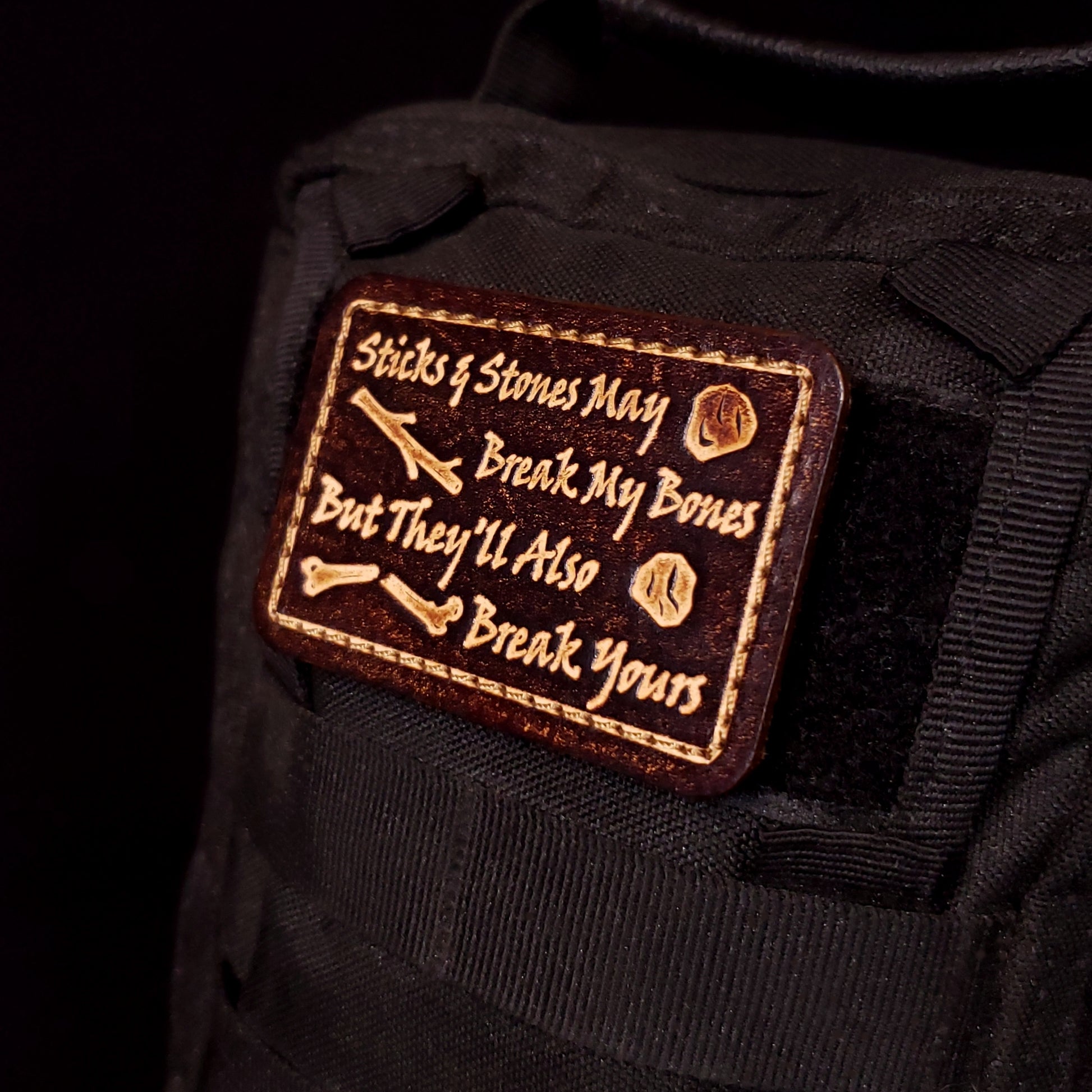 Brown rectangular velcro patch embossed with text and graphics of sticks, rocks, and a broken bone, affixed to a bag.