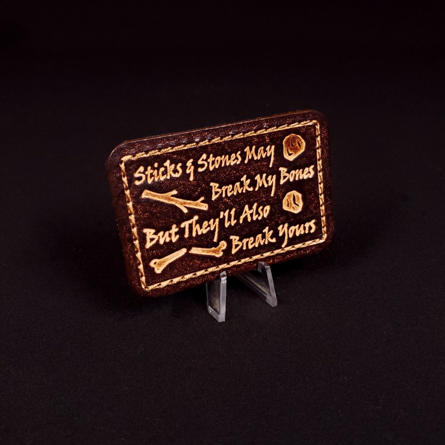 Brown rectangular velcro patch embossed with text and graphics of sticks, rocks, and a broken bone.
