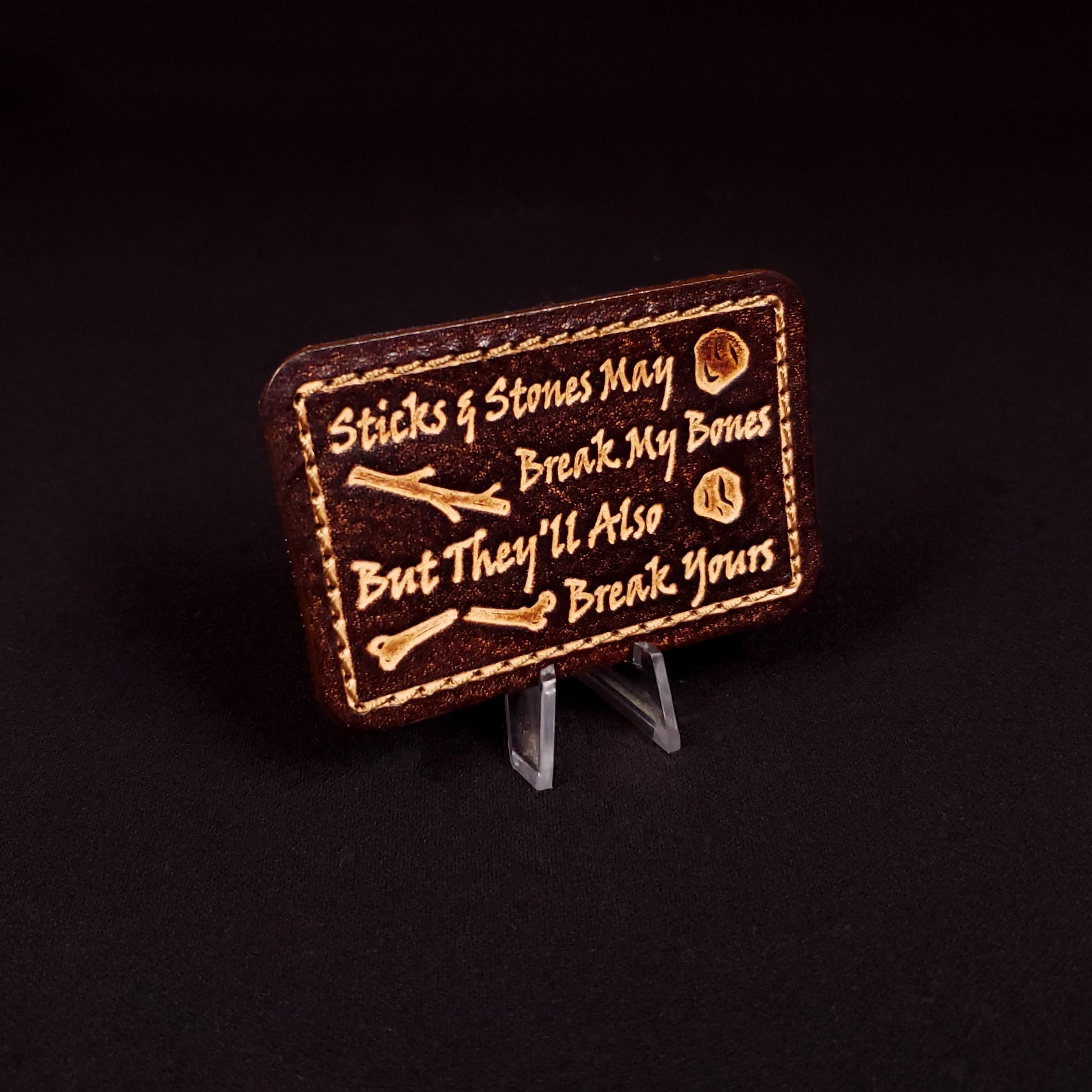 Brown rectangular velcro patch embossed with text and graphics of sticks, rocks, and a broken bone.