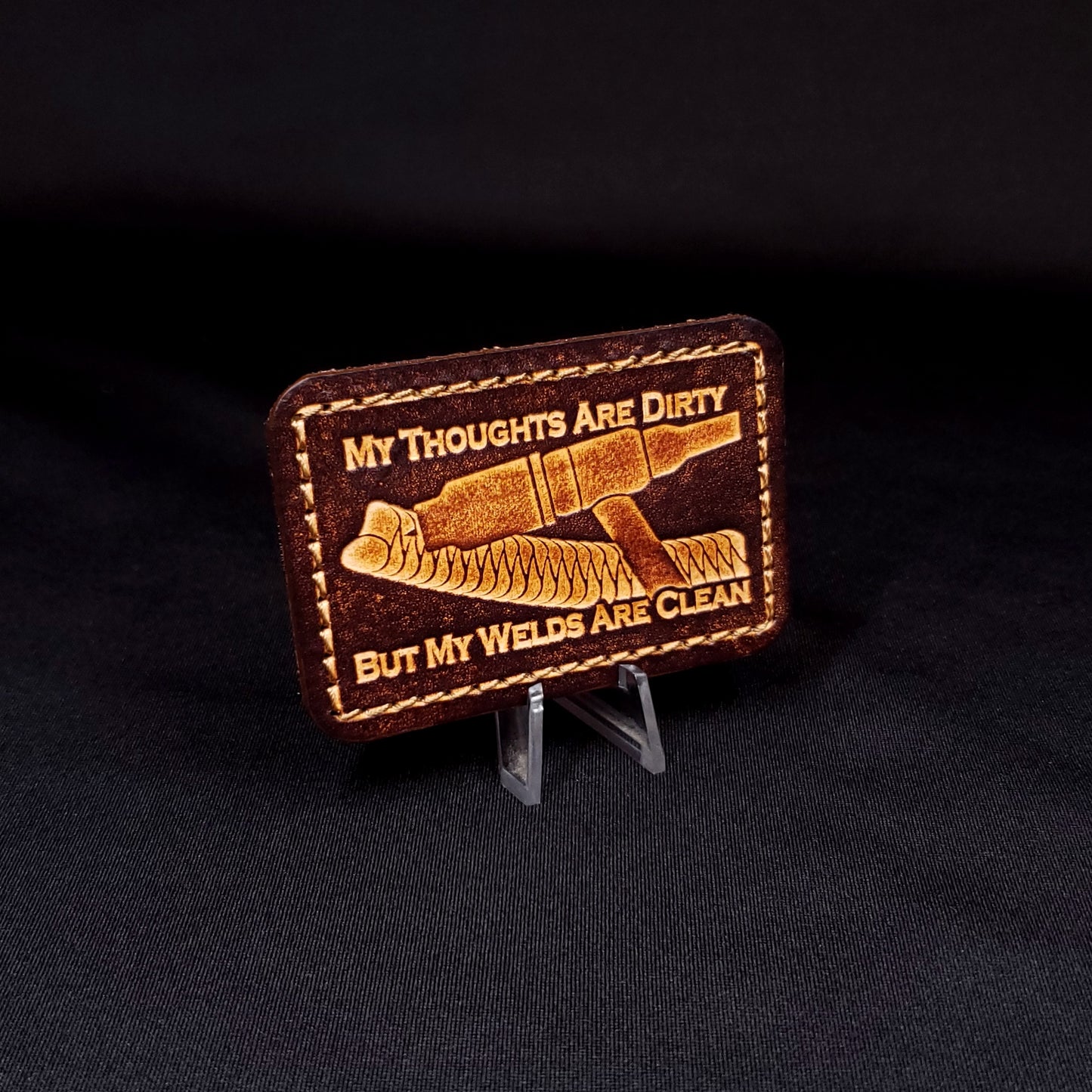 Brown rectangular velcro patch embossed with a picture of a welding tig torch and text, posed on a black background.