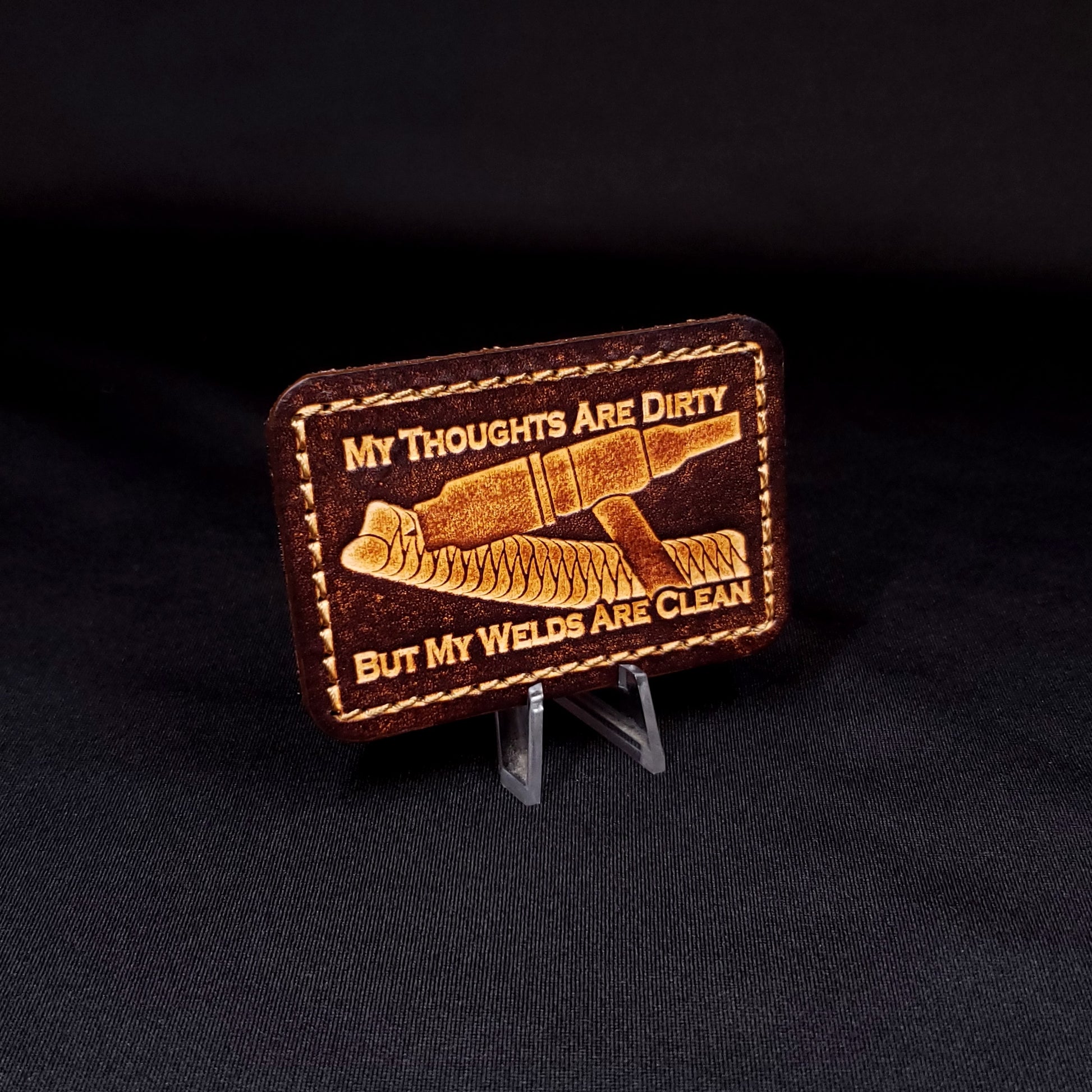 Brown rectangular velcro patch embossed with a picture of a welding tig torch and text, posed on a black background.