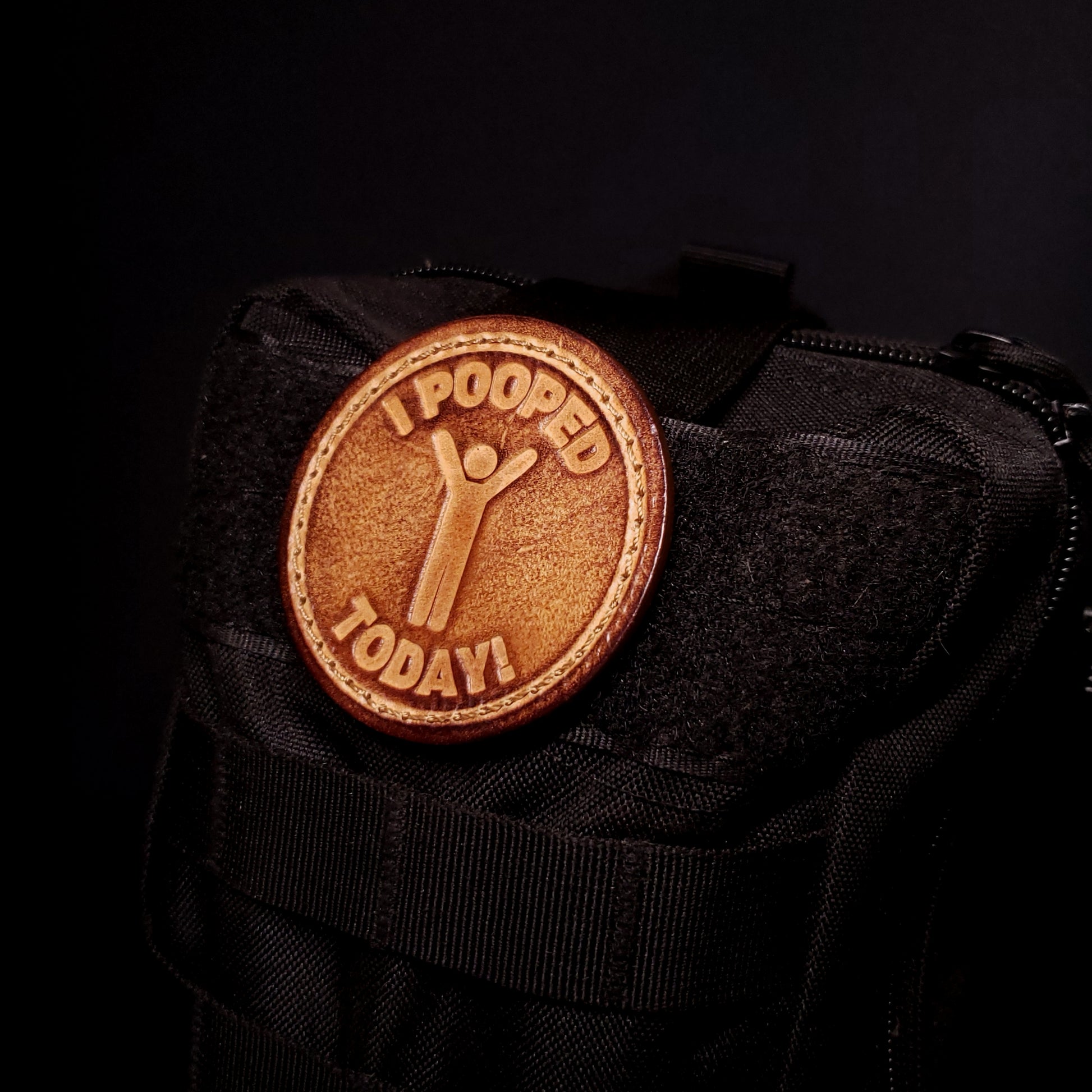 Brown leather velcro patch embossed with text that says "I pooped today", and a graphic of a cheerful person, affixed to a bag.