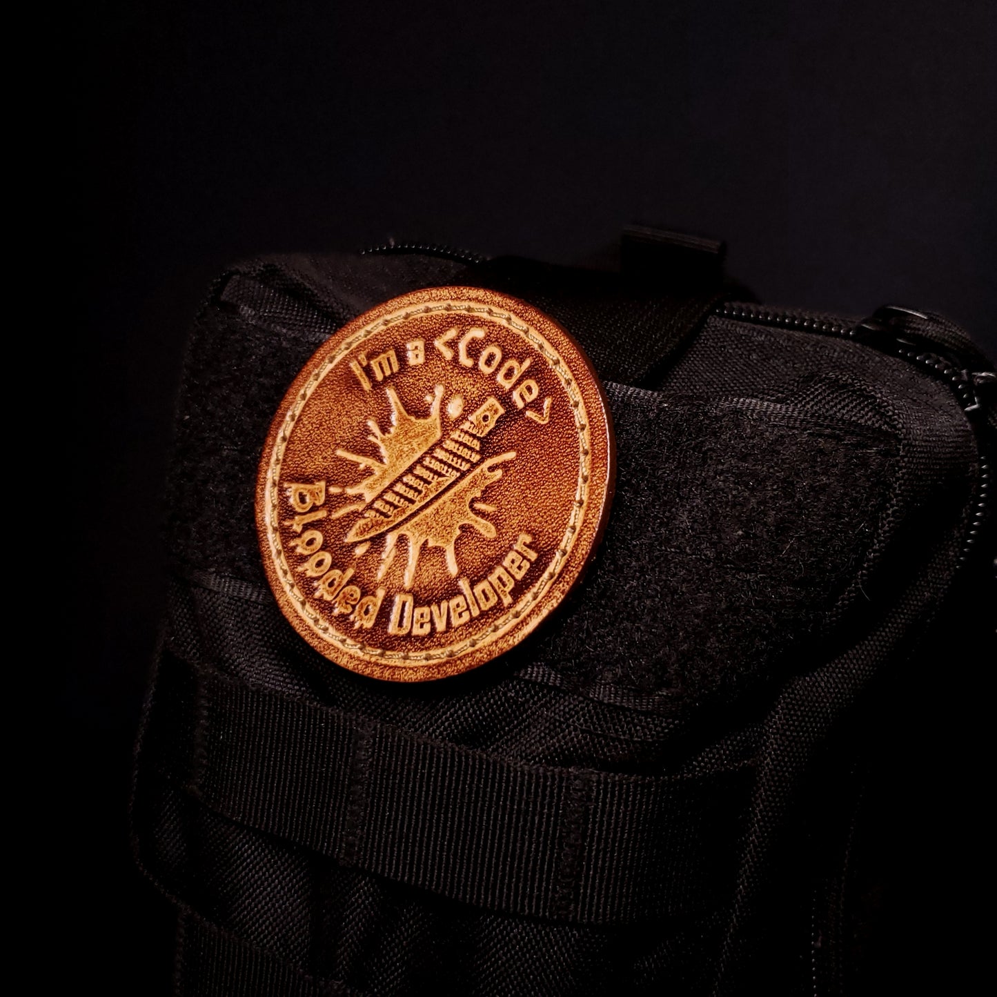 Brown leatehr velcro patch featuring text and a graphic of a knife made of ones and zeros on a blood splatter, affixed to a bag.