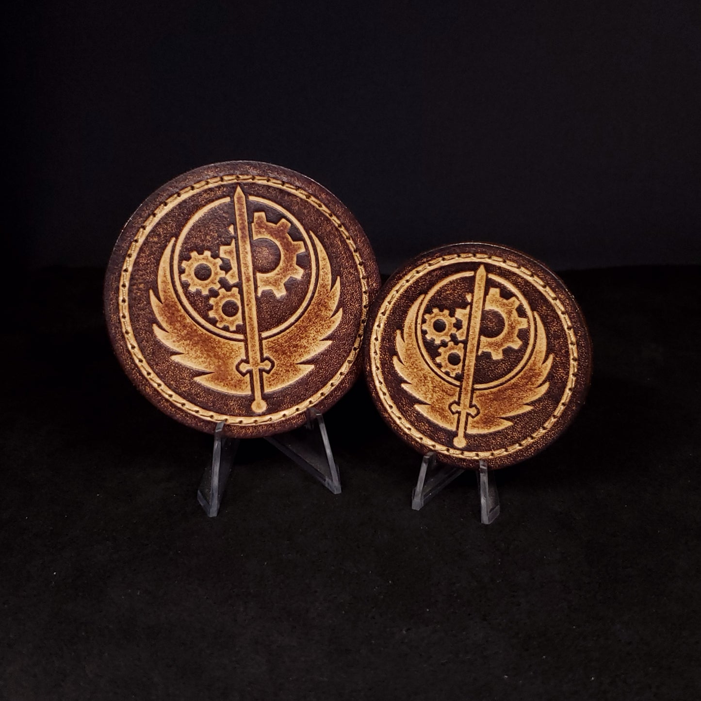 Two brown circular velcro patches embossed with the Brotherhood of Steel emblem, posed on a black background.