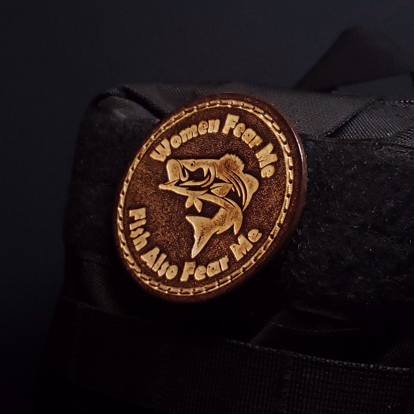 Brown circular velcro patch embossed with text and a graphic of a fish, affixed to a bag.