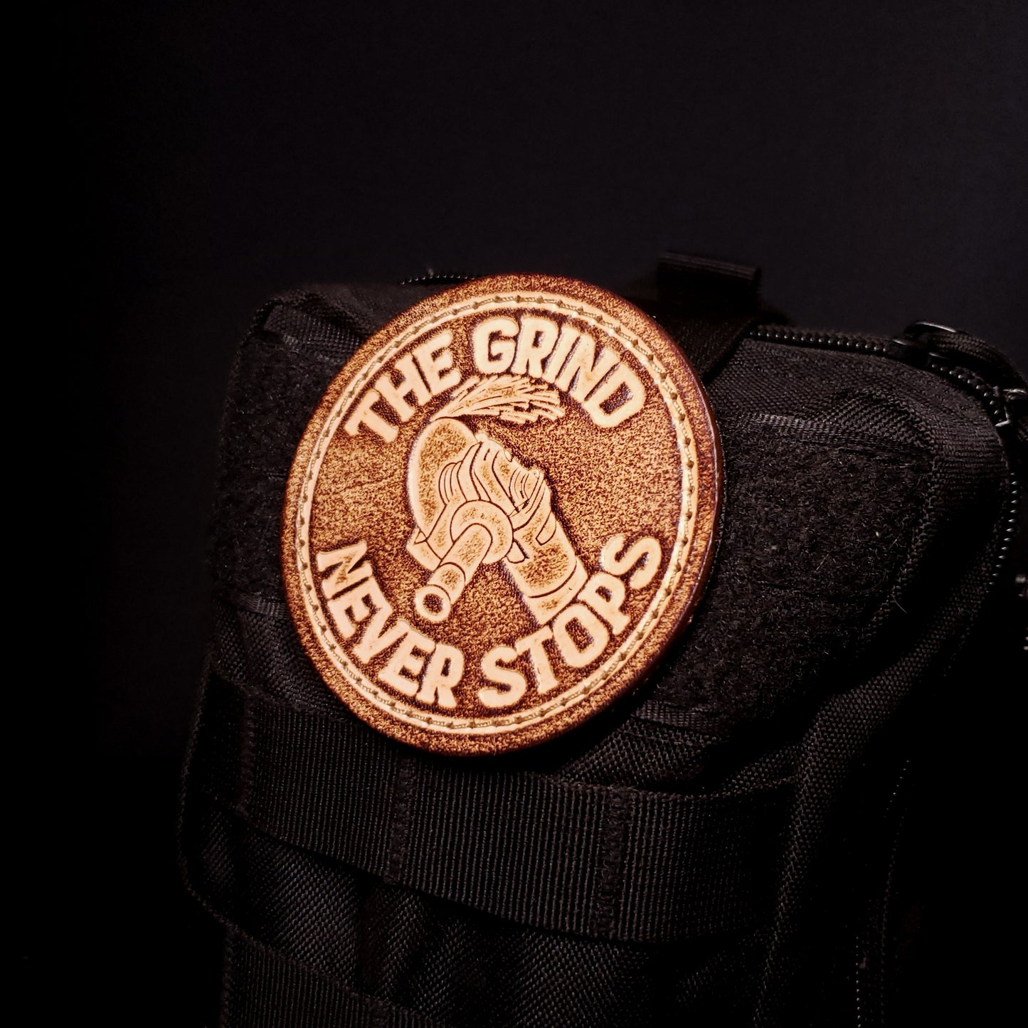 Brown leather velcro patch with text that says "The grind never stops" and an image of an angle grinder, affixed to a bag.
